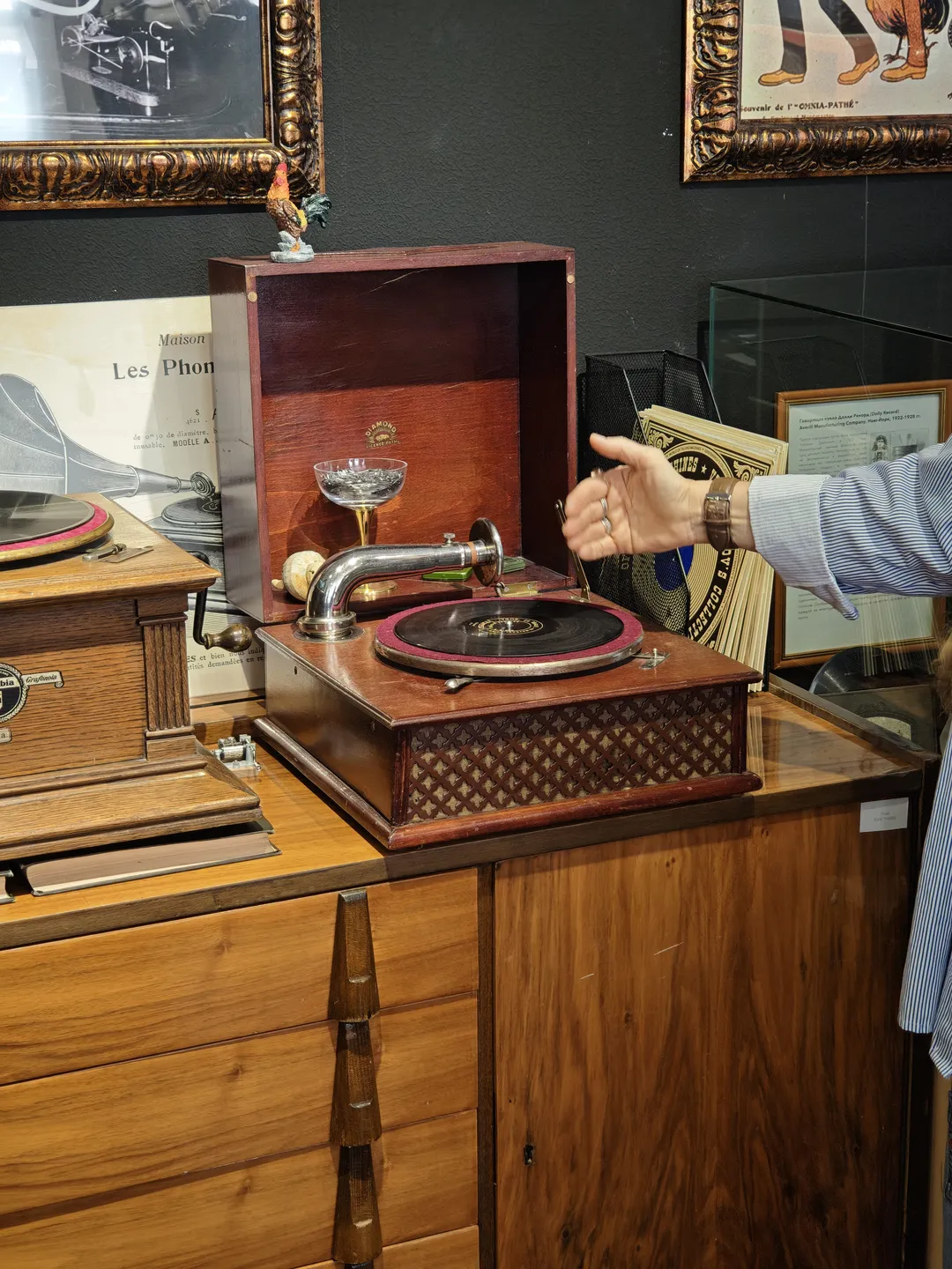 A gramophone with a wooden casing placed on a cabinet, with a hand operating it. The gramophone has a retro design and features a metal horn, with other similar equipment and decorations in the background.