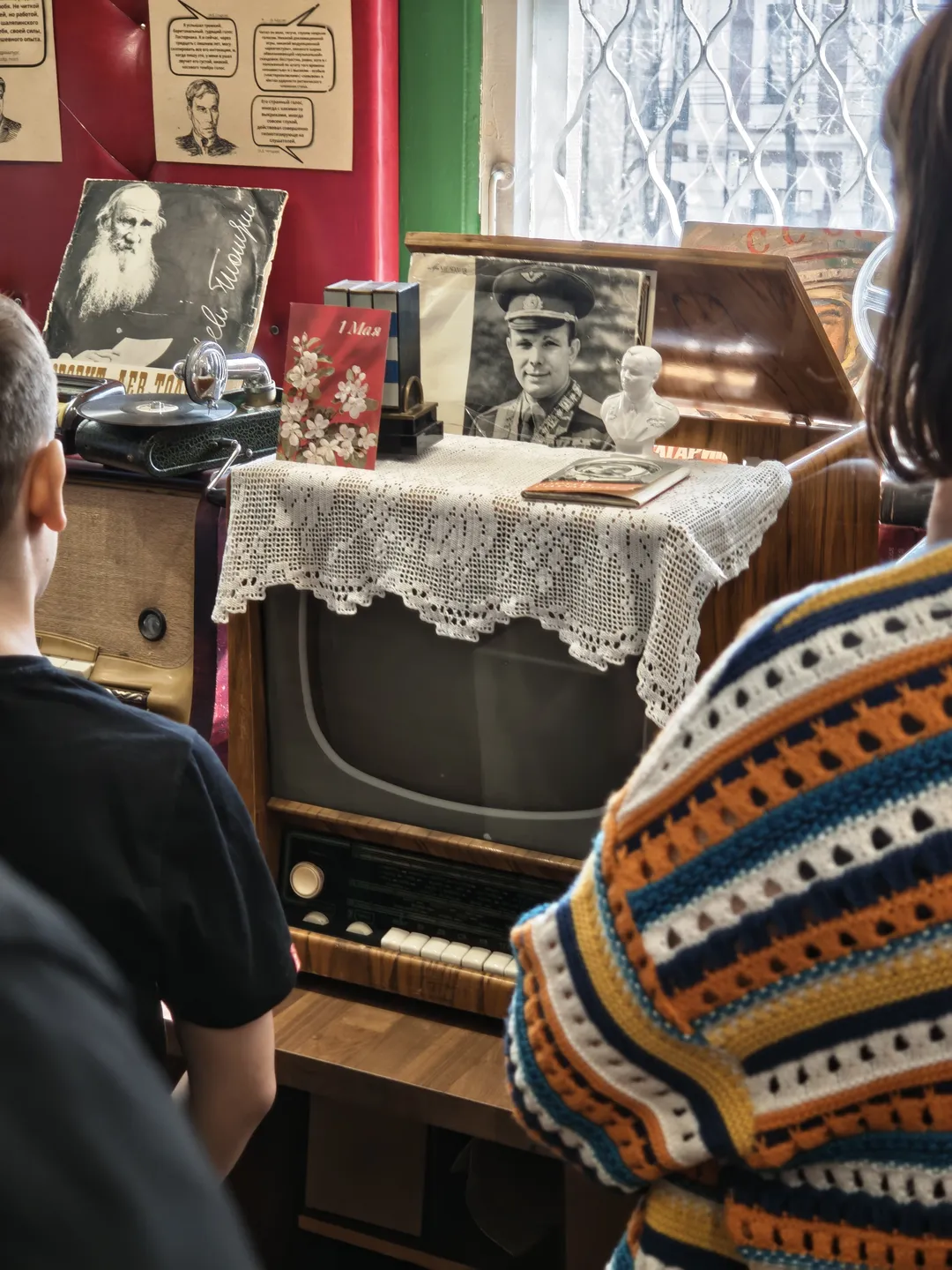 Two people standing in front of a vintage television set covered with a white lace cloth. Two black-and-white photos hang above the TV next to a small figurine, against a red wall adorned with some posters.