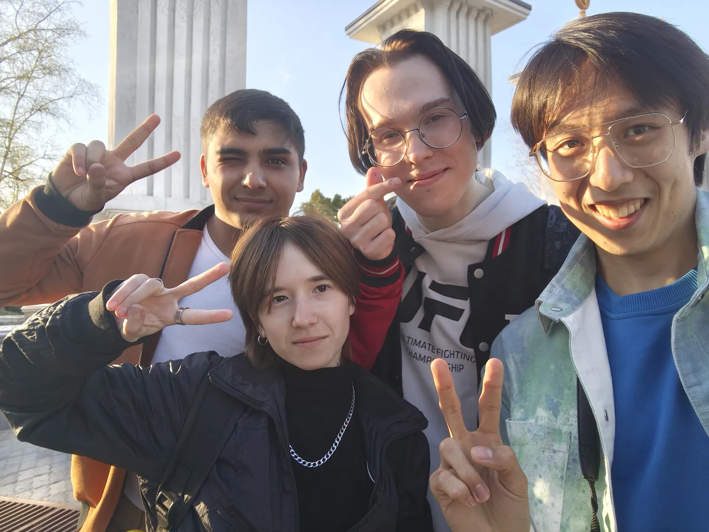 Four people posing together for a group photo, with three of them flashing peace signs, against a background of pillars and the sky.