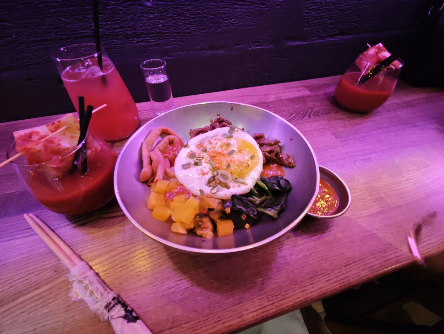 A bowl of food containing fried eggs, vegetables, and meat on a table, with several glasses of drinks nearby, one of which has a straw. Chopsticks are placed on the bottom-left corner of the table.