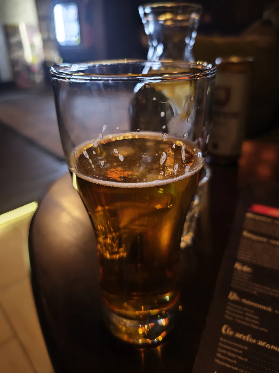 A glass of golden beer with foam on top placed on a tabletop. Menus and other tableware are visible in the background, and the glass reflects the surrounding light.
