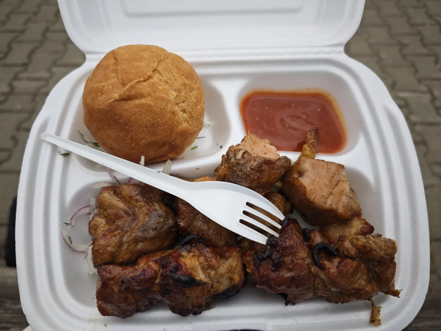 Chunks of grilled meat, a small bun, and a red sauce served in a white foam container, with a white plastic fork placed on top.