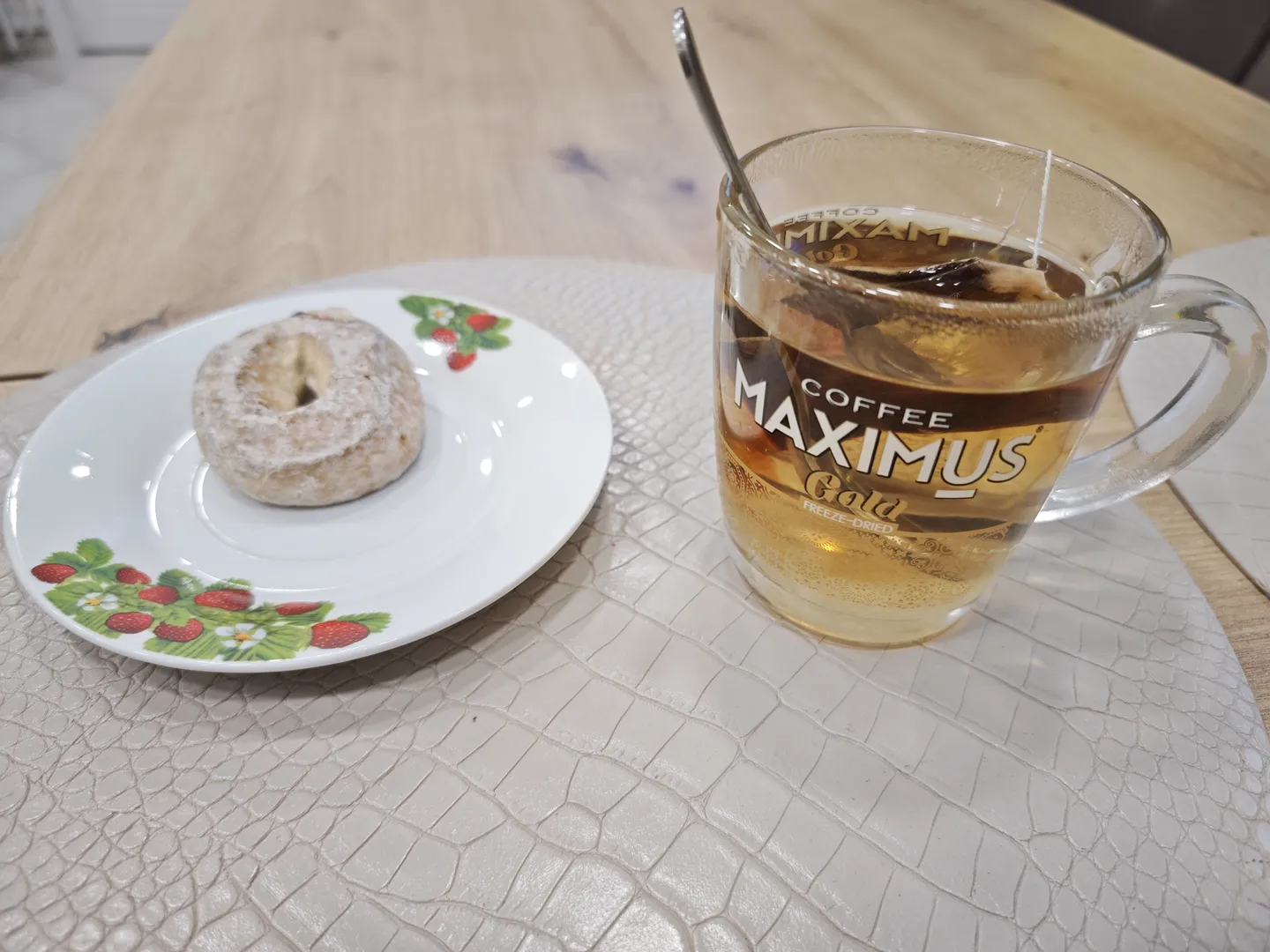 A transparent glass cup and a white plate placed on a table with a crocodile skin texture. The cup contains tea and is printed with the text 'COFFEE MAXIMUS Gold FREEZE-DRIED', while the plate holds a doughnut dusted with powdered sugar.