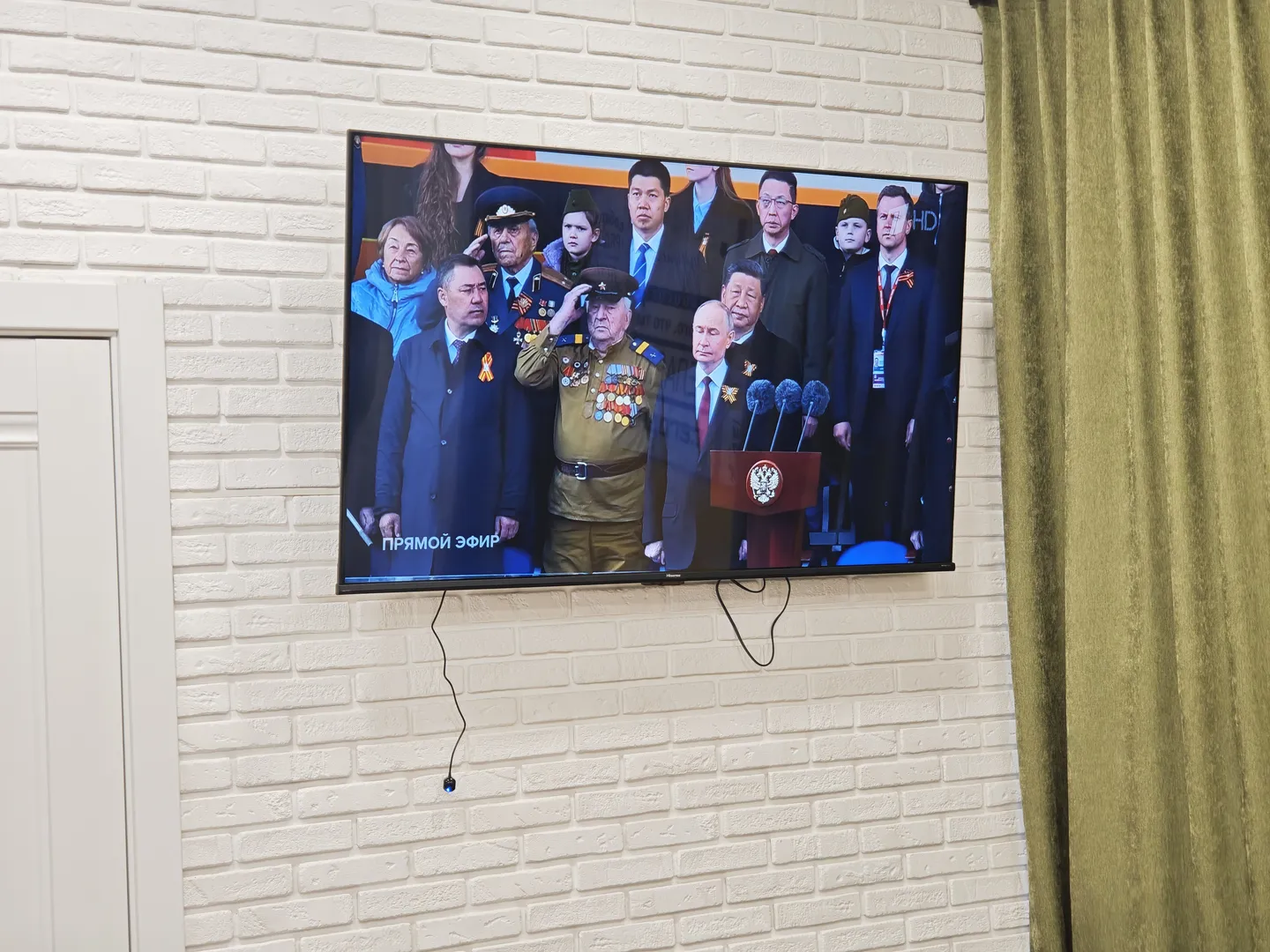 A hotel television broadcasting the military parade celebrating the 80th anniversary of the victory in the Great Patriotic War, showing a group photo of national leaders on screen.