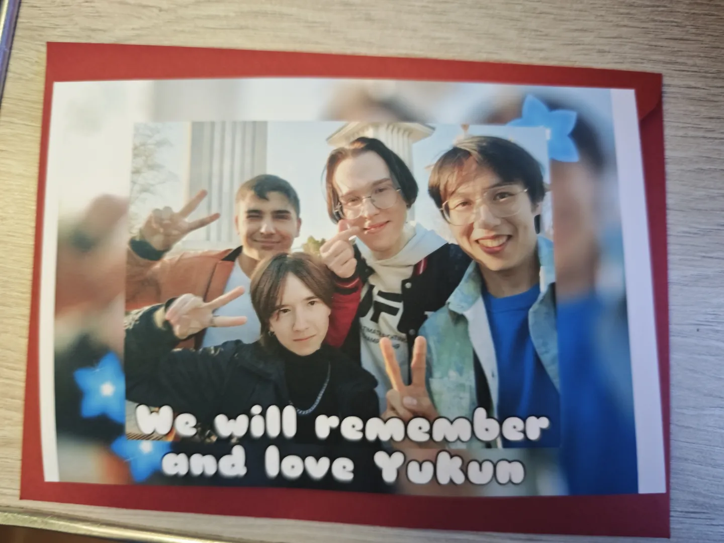A photograph featuring four people all flashing peace signs, with the text 'We will remember and love Yukun' written below. The photograph is decorated with a red border and blue star patterns, with part of a building structure visible in the background.
