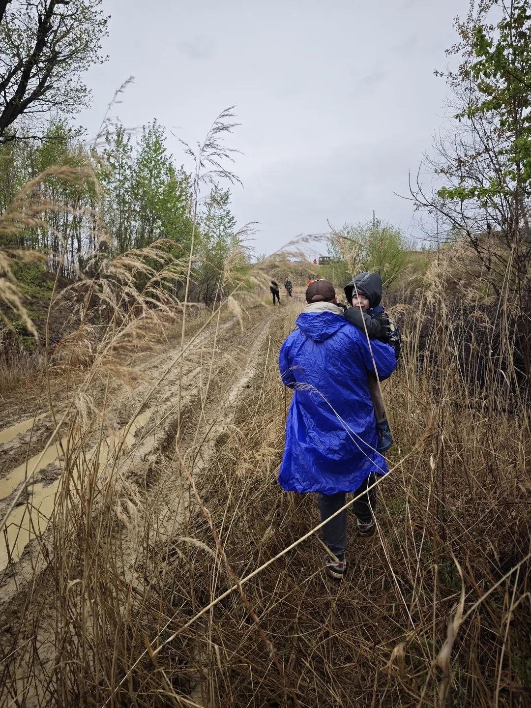 Two people wearing blue raincoats walking on a lawn, surrounded by tall dry grass. A few people are active on the grass in the distance, with trees and a vehicle in the background under an overcast sky.