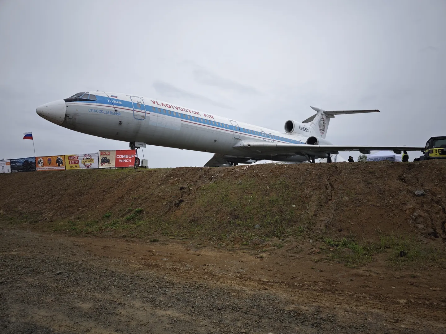 A large airplane parked on a dirt slope, with the fuselage marked with 'VLADIVOSTOK AIR'. Flags and billboards stand in front of the airplane, with some trees in the background on the relatively flat dirt ground.