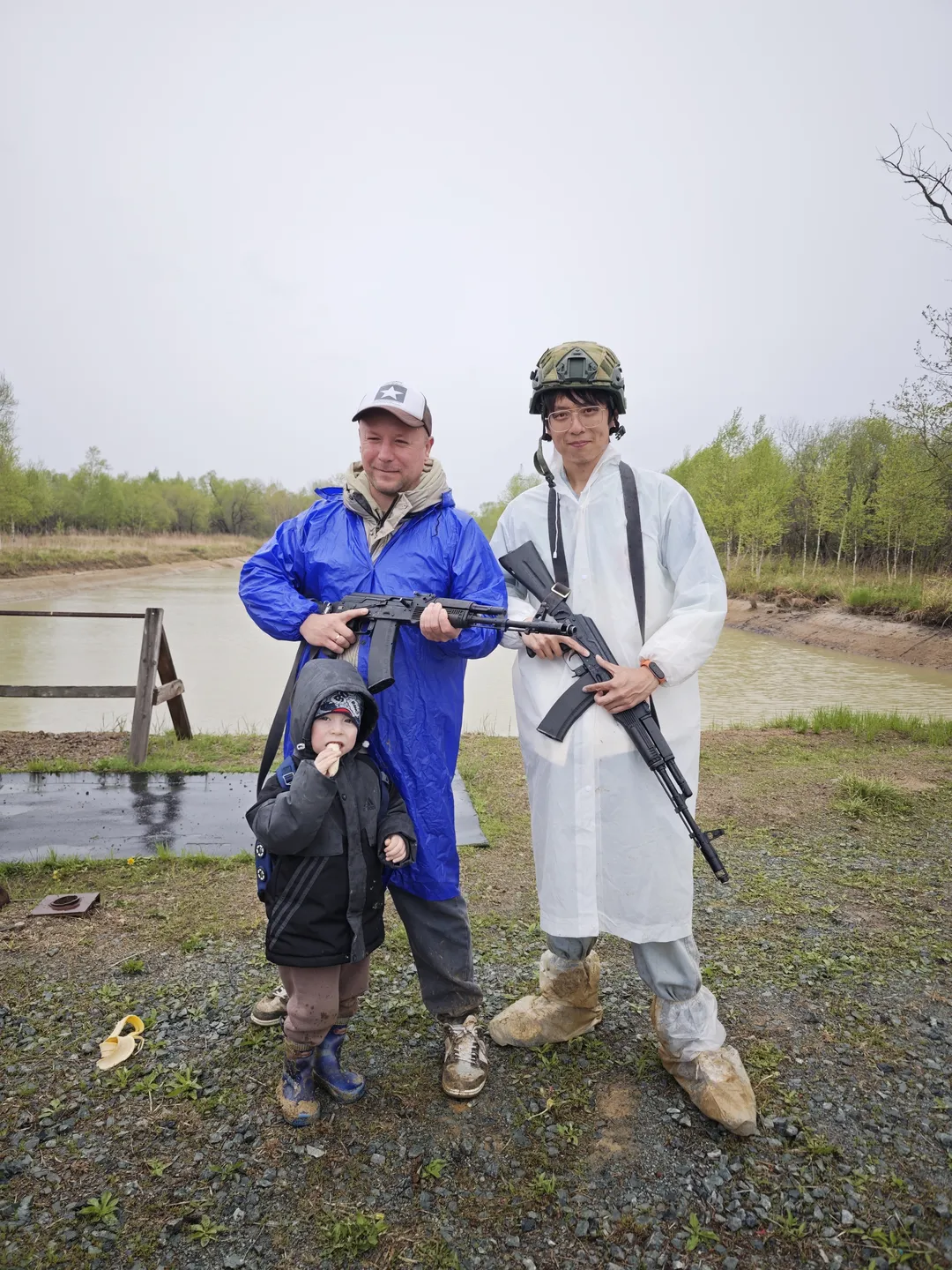 Two adults and a child standing together, each holding a gun. The person on the left wears a blue coat, and the person on the right wears a white coat and a helmet, against a background of a river and trees.