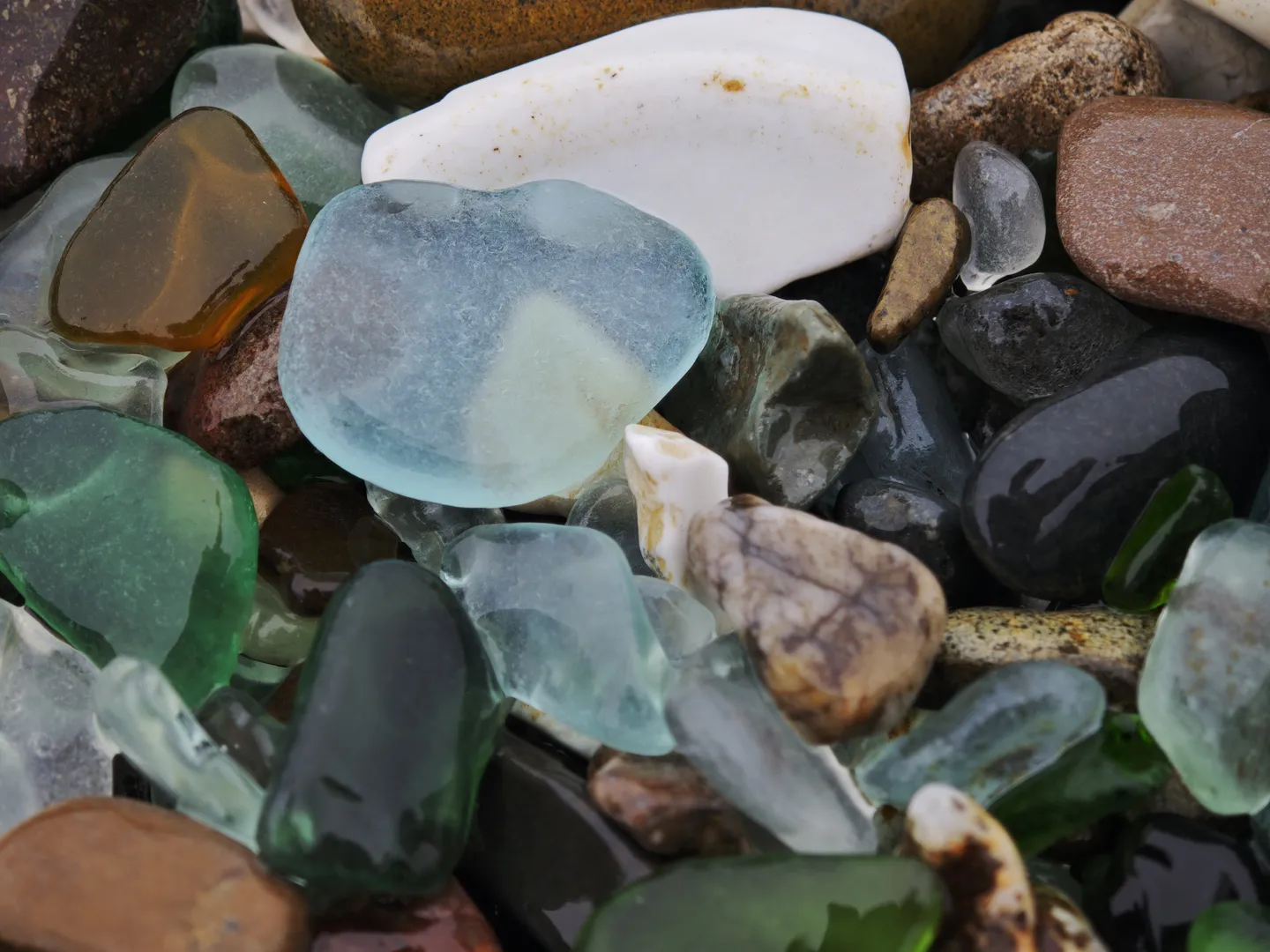 A pile of glass fragments and pebbles in rich colors, including blue, green, brown, and transparent. The fragments are irregularly shaped with smooth surfaces, mixed together with the pebbles.