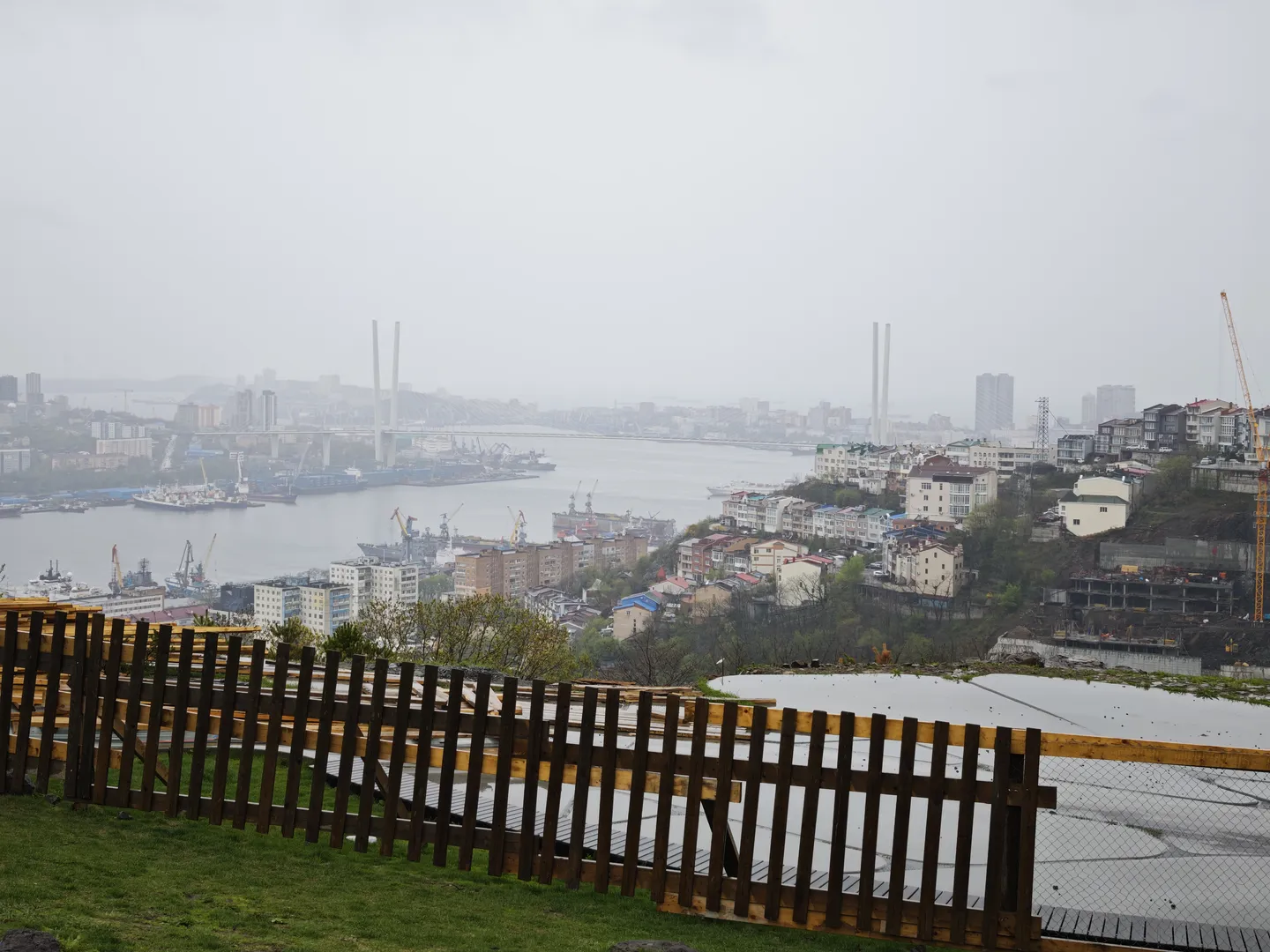 A bird's-eye view of a cityscape, with a wooden fence in the foreground and a dense cluster of buildings and a river in the background. Bridges span across the river, and both shores are lined with high-rise buildings and cranes under a cloudy sky.