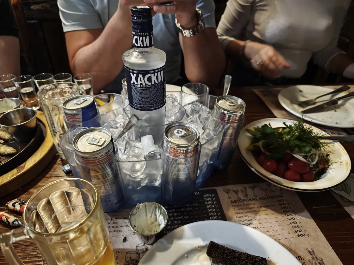 A variety of drinks and food arranged on a dining table, with a bottle labeled 'ХАСКИ' in the center, flanked by canned drinks and a bowl of salad. Empty glasses and ice cubes are on the table, and several people sit around with various hand gestures.