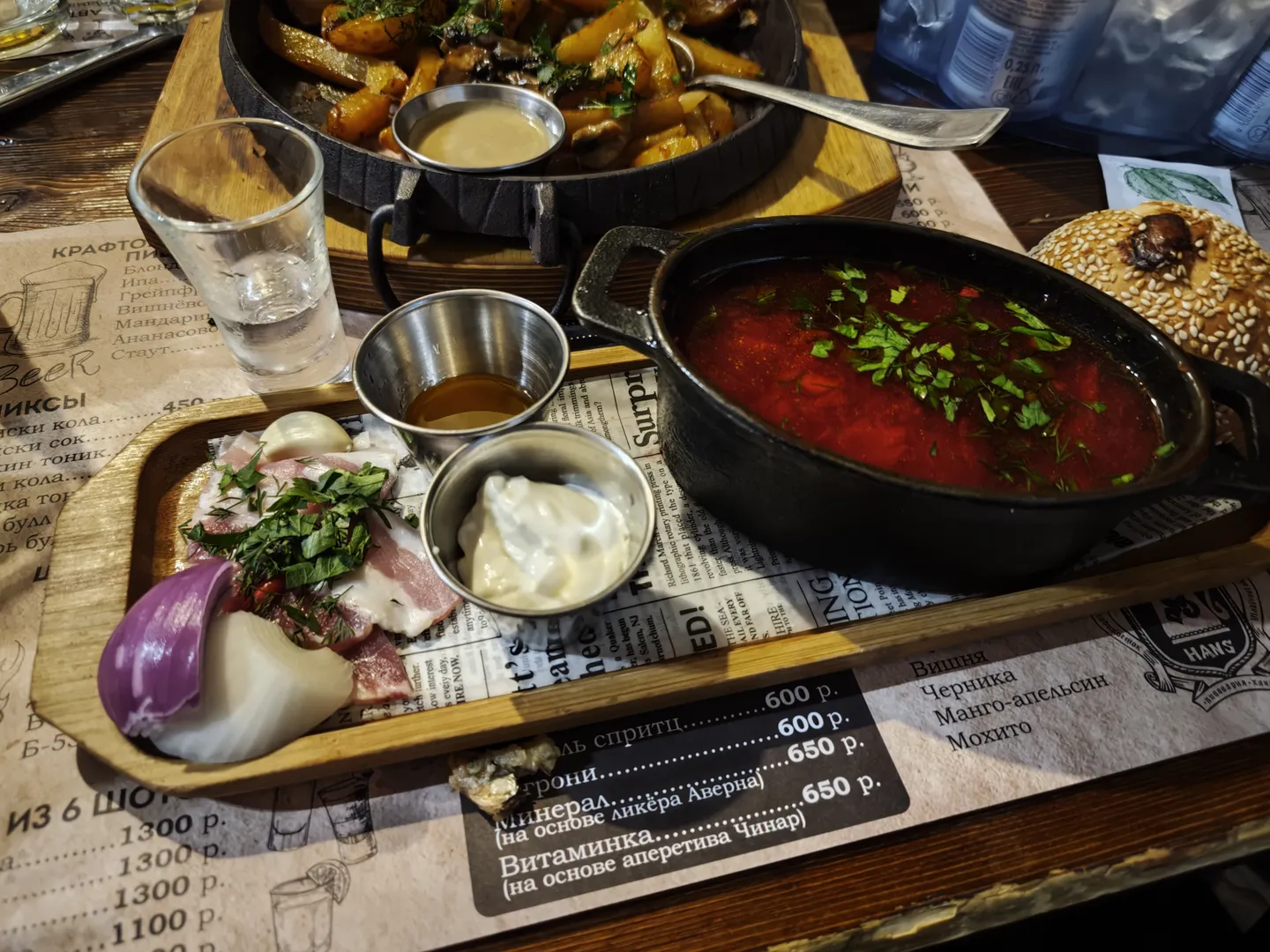 A pot of red soup served in a black pot on a dining table, accompanied by sliced onions and seasonings, with side ingredients in small bowls nearby. A paper menu lists various drink prices.