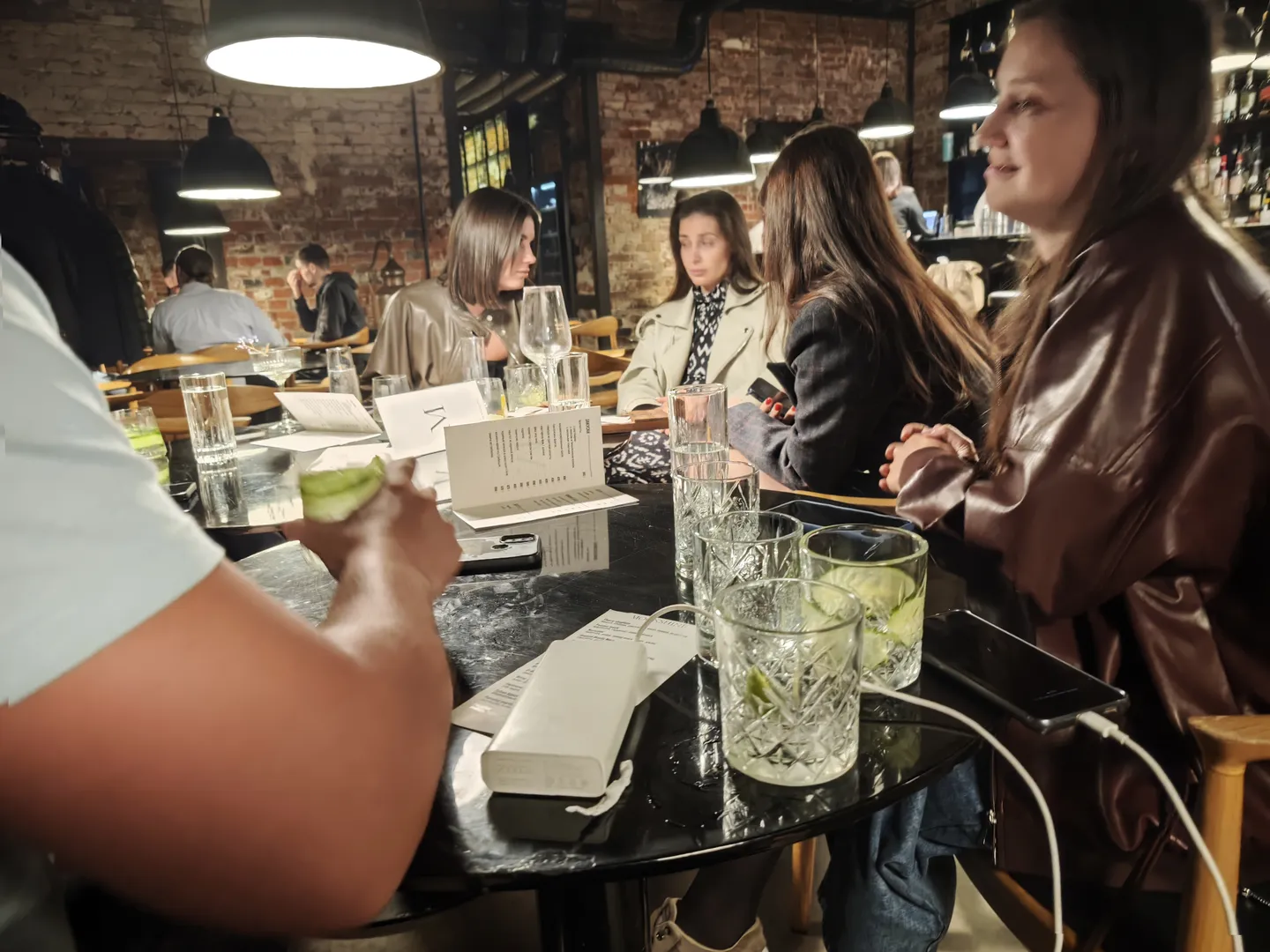 Multiple people gathered around a dining table inside a restaurant, with transparent glasses and menus placed on the table. A man is putting a lemon slice into a glass while several women sit opposite him, within a modern interior under warm lighting.