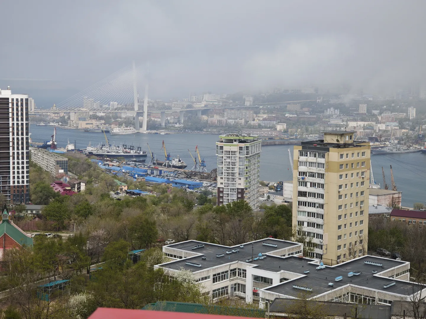 A cityscape with a balanced mix of high-rise buildings and low-rise structures, with a river in the middle featuring bridges and vessels. The distant buildings are shrouded in thin mist under an overcast sky, with green vegetation along both shores of the river.
