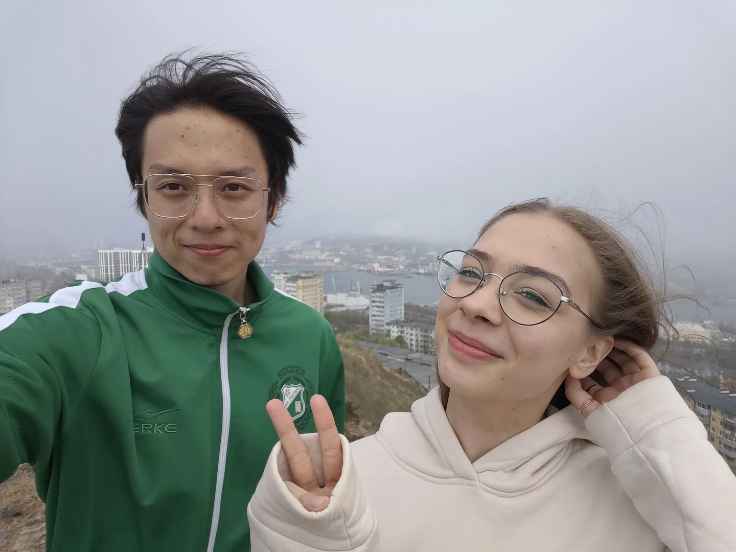 Two people smiling and taking a selfie from a high vantage point, with the cityscape and harbor in the background. The person on the left wears a green jacket, and the person on the right wears a white hoodie.