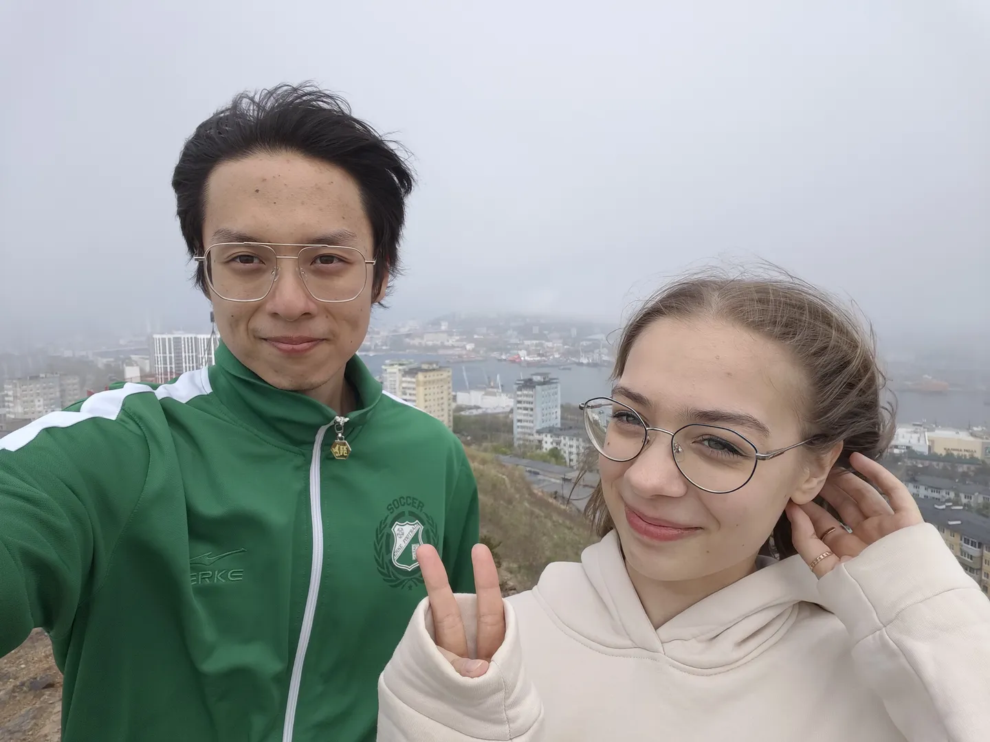 Two people smiling and taking a selfie from a high vantage point, with the cityscape and harbor in the background. The person on the left wears a green jacket, and the person on the right wears a white hoodie.