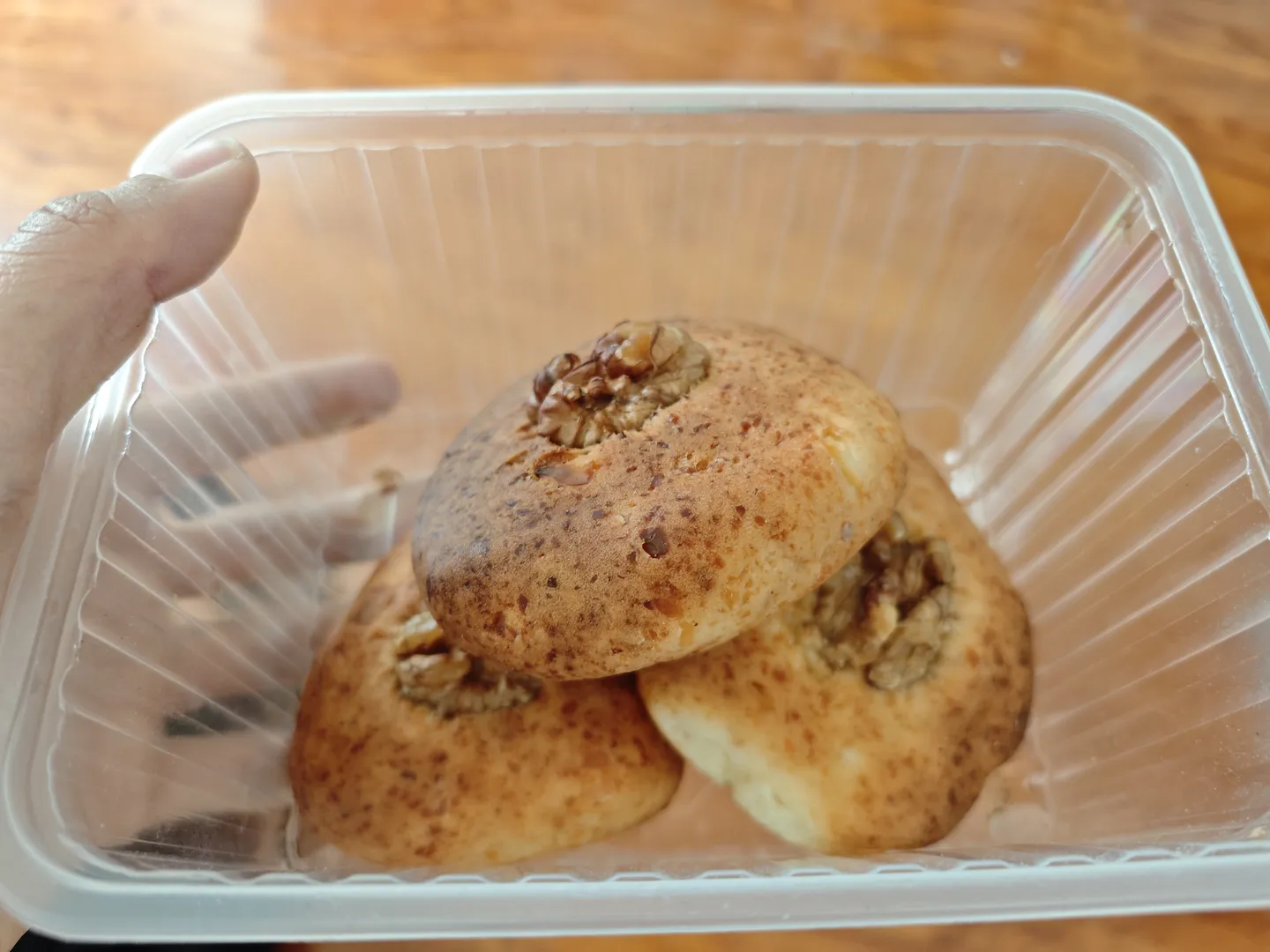 A transparent plastic container holding three round pastries topped with crushed nuts. The pastries are golden brown and regularly shaped, with a hand holding the plastic container.