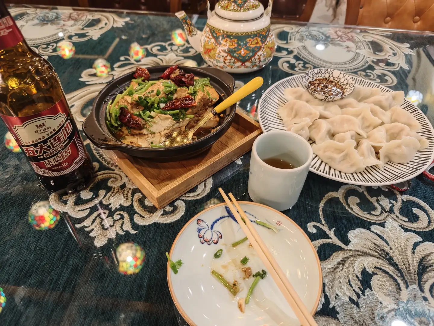 餐桌上摆放着一盘饺子、一锅热菜、一瓶啤酒和一杯茶。热菜盛在黑色砂锅中,上面有青椒和干辣椒,旁边放着一把黄色手柄的勺子。桌布是深绿色带花纹的,中央有一只空盘子,上面留有食物残渣和一双筷子。