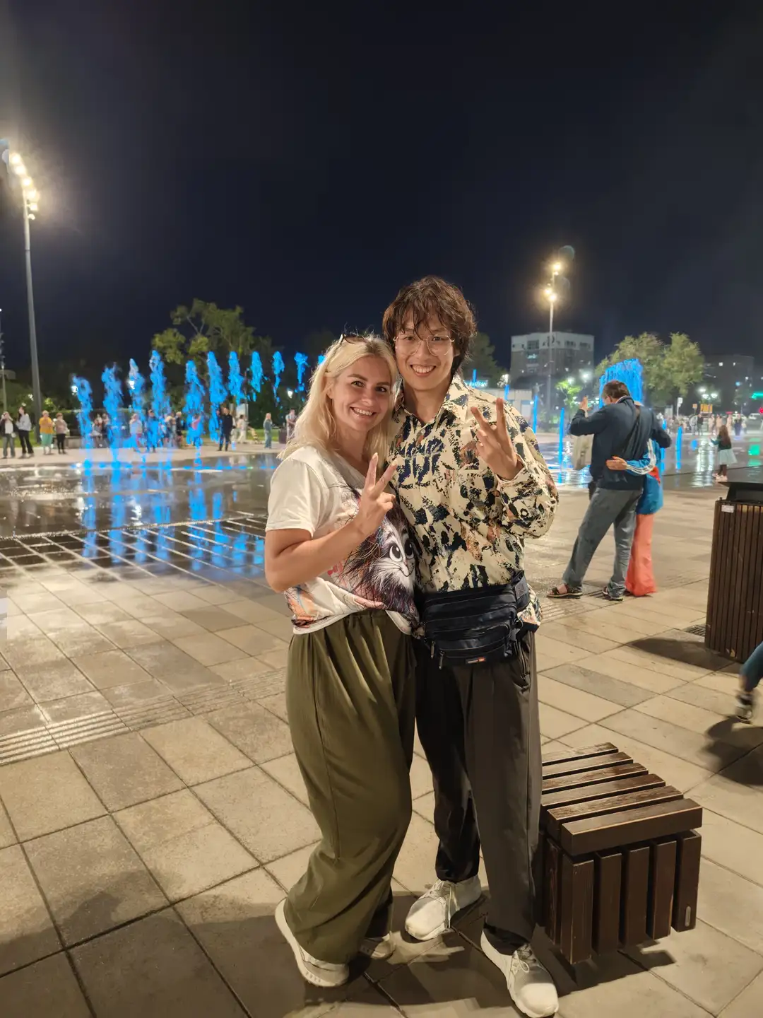 两个人在夜晚的广场上合影,背景是蓝色灯光喷泉和一些行人。左边的人穿着白色T恤和绿色宽松裤子,右边的人穿着图案衬衫和黑色裤子,腰间挎着一个包。两人都面带微笑,比出胜利手势。