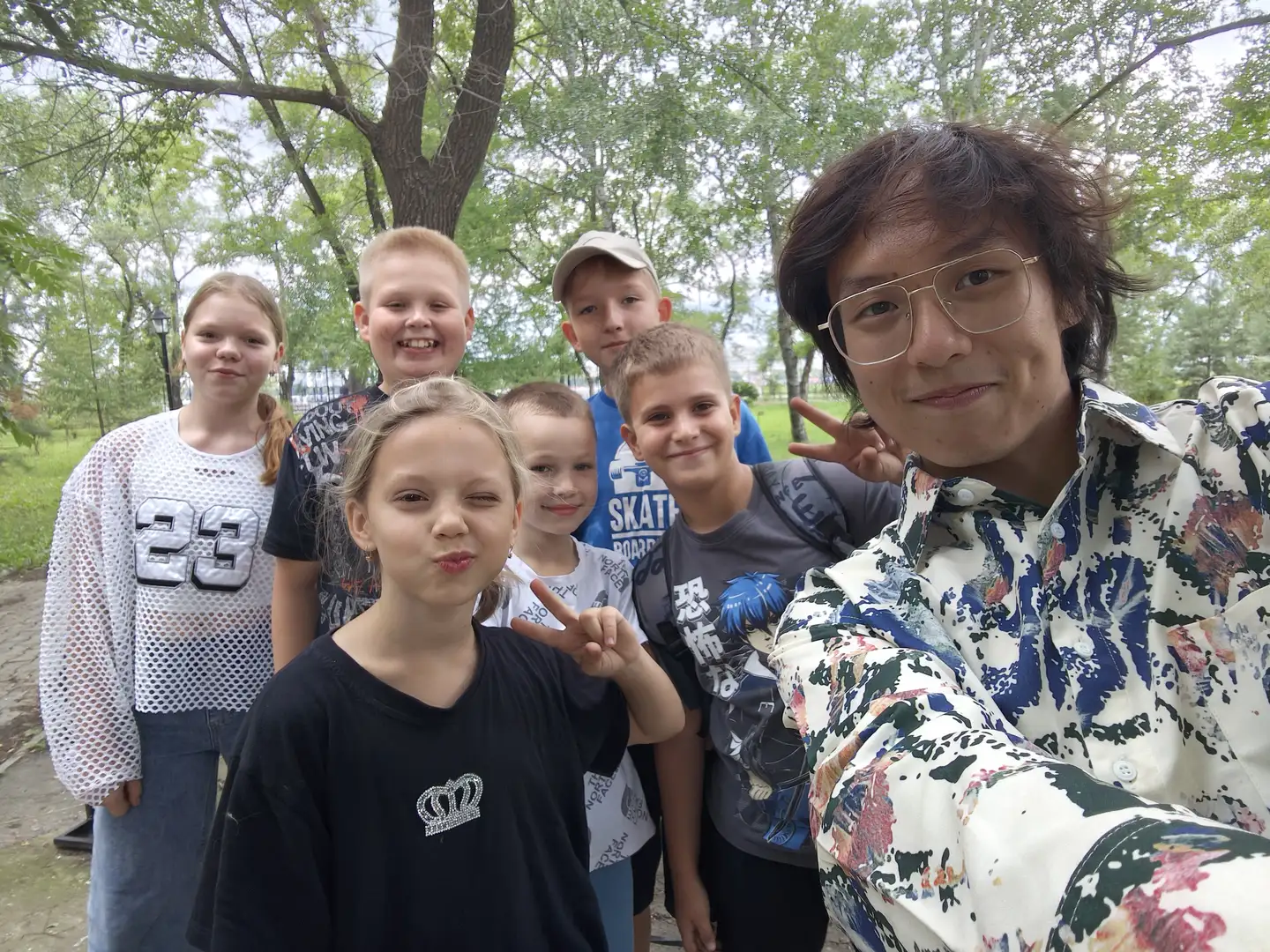 一群孩子和一名戴眼镜的成年人在户外合影,背景是树木和公园小径。孩子们穿着休闲服装,有的比出胜利手势,表情自然。成年人身穿花纹衬衫,站在最右侧。