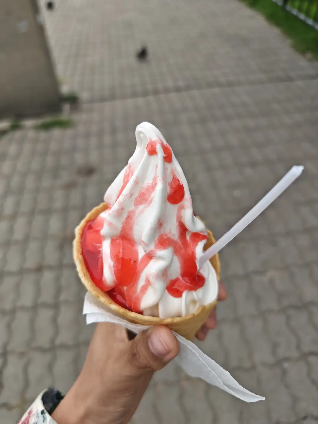 一只手拿着一个冰淇淋蛋筒,里面装着白色软冰淇淋,上面淋有红色果酱,蛋筒用纸包裹,插着一根塑料勺子