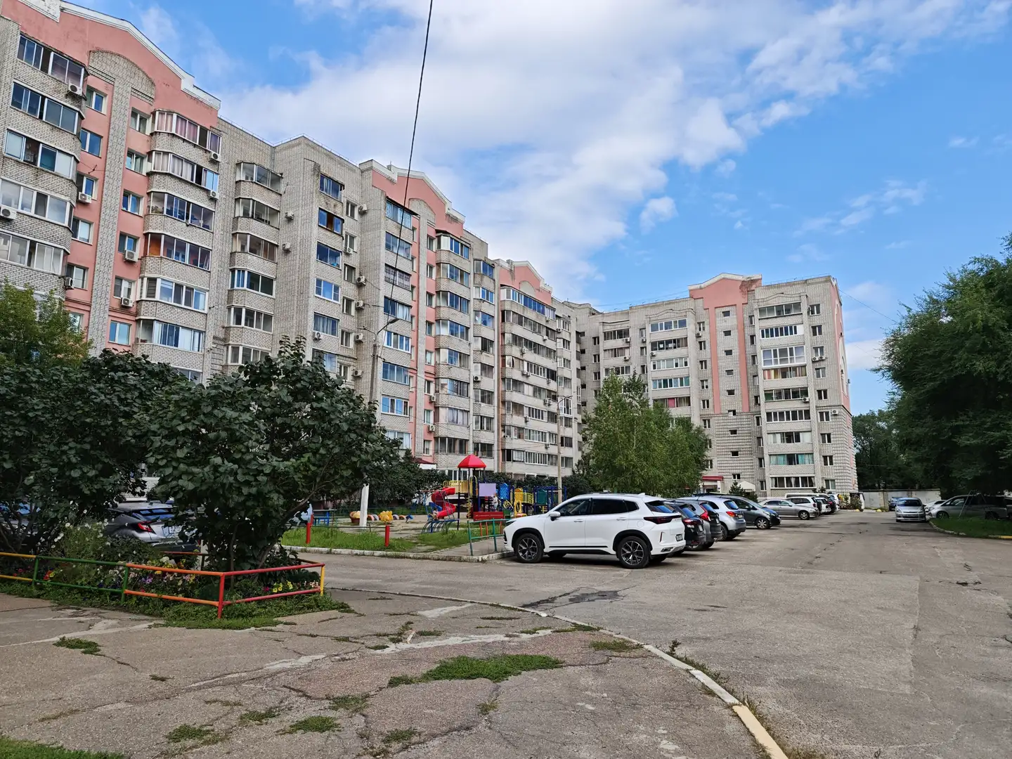 一排高层住宅楼,外立面为灰白与粉红相间,楼前有停车场和绿化带。天空晴朗,云朵稀疏,楼间可见儿童游乐设施和停放的车辆。
