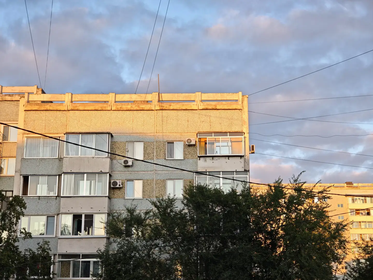 一栋多层住宅楼的顶部在夕阳照射下泛着金黄色光芒,窗户排列整齐,部分装有空调外机。建筑外墙为浅色与深色相间,屋顶边缘有护栏结构。天空中布满灰蓝色云朵,电线横跨画面。
