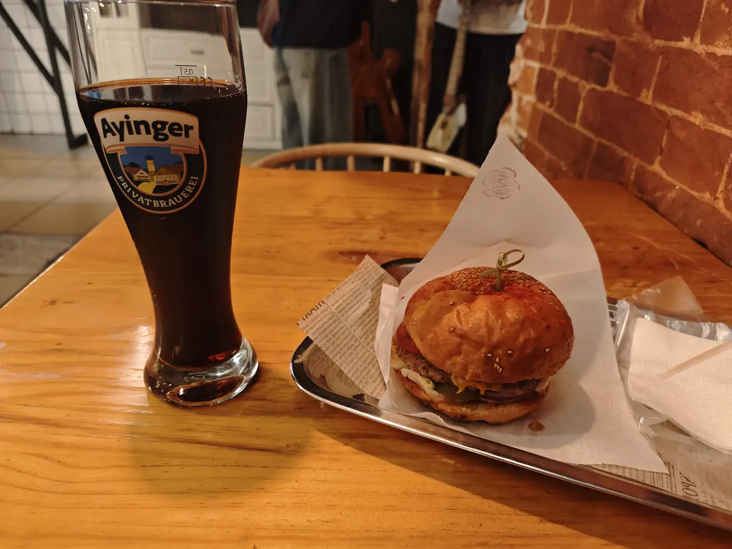 木桌上摆放着一杯深色啤酒和一个汉堡,啤酒杯上有“Ayinger PRIVATBRAUEREI”的标志。汉堡放在金属托盘上的纸巾中,夹有生菜、番茄和芝士,用牙签固定。背景可见砖墙和模糊的人影,环境像是餐厅或酒吧内部。