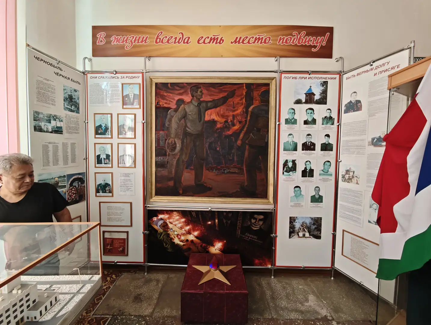展览墙上有大幅画作和多张照片,中央画作描绘了两名男子在火场中指挥救援的场景。上方横幅写着“В жизни всегда есть место подвигу!”,两侧展板展示了人物肖像和文字介绍。前方有一个红色盒子,上面放置着一颗金色五角星,旁边有玻璃展柜和一面三色旗。