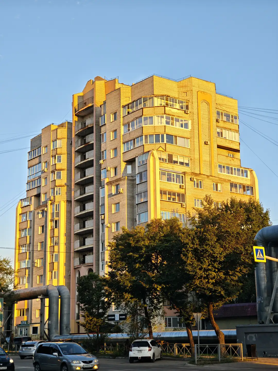 一栋高层住宅楼在夕阳下闪耀着金黄色的光芒,外立面由瓷砖和玻璃窗组成。楼体有多层阳台,窗户排列有序。前景中有几棵树和一辆行驶中的汽车,右侧有一个行人过街标志。