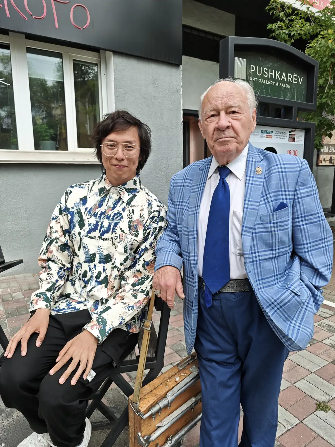 一位年轻男子坐在椅子上,旁边站着一位年长的男士,两人站在一家名为“PUSHKAREV ART GALLERY & SALON”的画廊前。年轻男子穿着印花衬衫和黑色裤子,年长者穿着蓝白格纹西装外套和蓝色领带。背景中可见绿植和部分建筑细节。