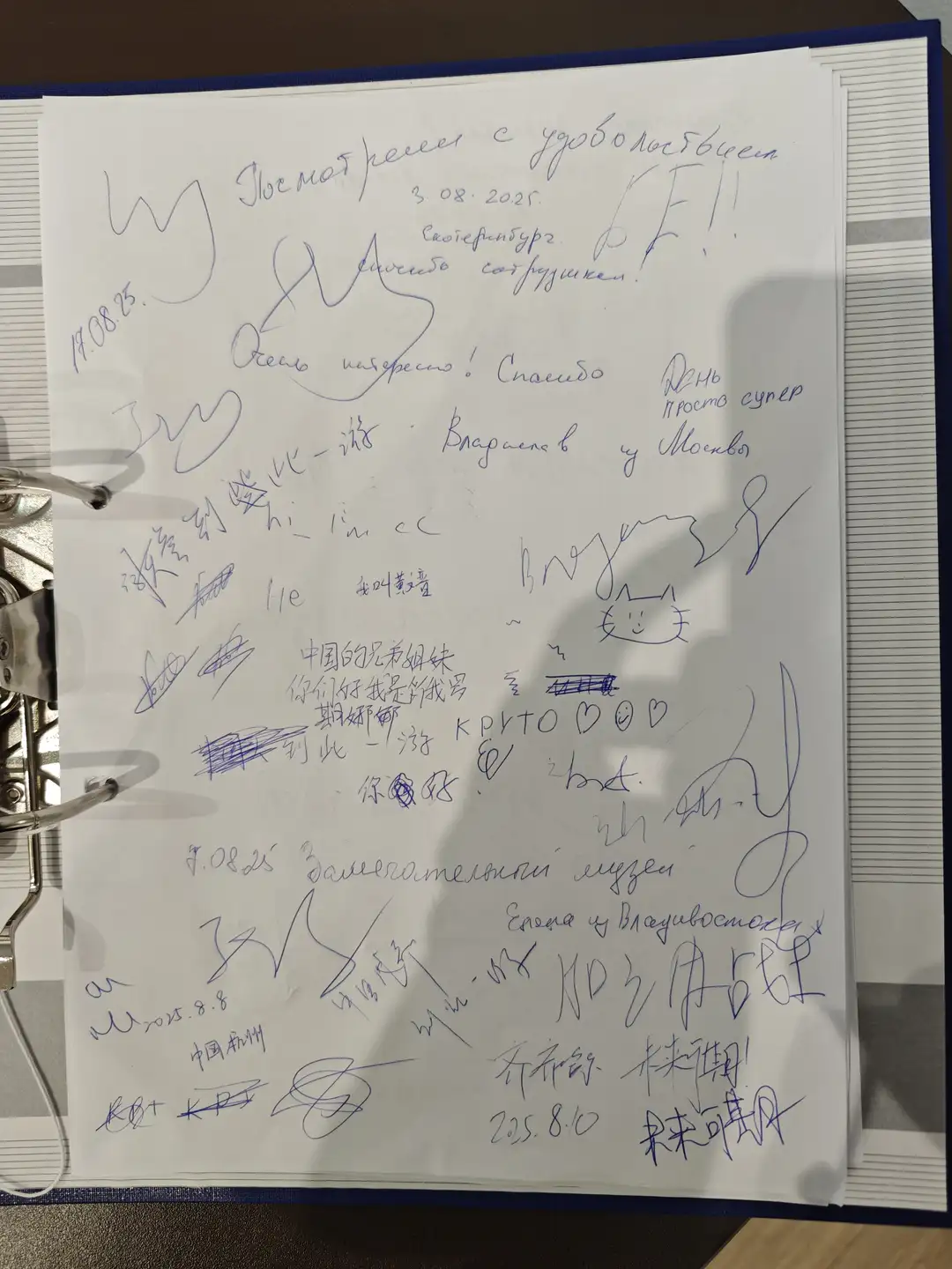 一本留言簿的一页,上面写满了不同语言的签名和留言,包括俄文、中文和英文。内容表达了对展览的兴趣和感谢,有的还画了笑脸或心形符号。日期显示为2025年8月,来自中国杭州、莫斯科等地的游客留下印记。