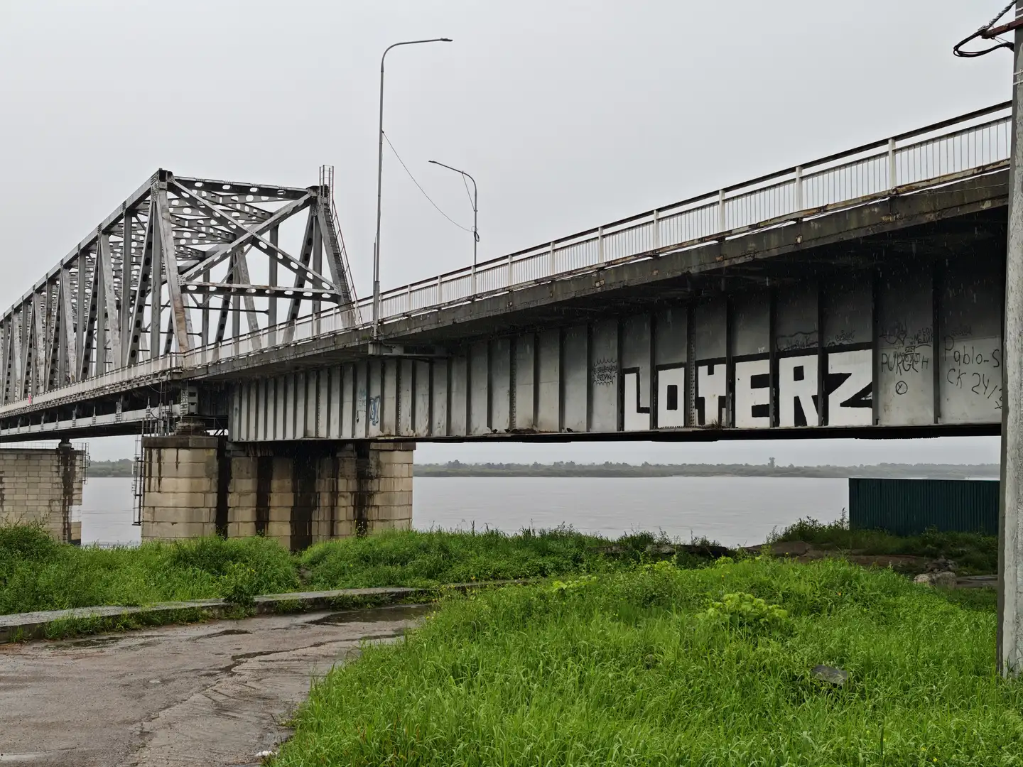 一座桥梁的侧面涂有“LOHERZ”字样,桥下是绿色的草地和一条小路。桥体由金属构成,支撑柱为混凝土。远处是宽阔的水面和对岸的景物。