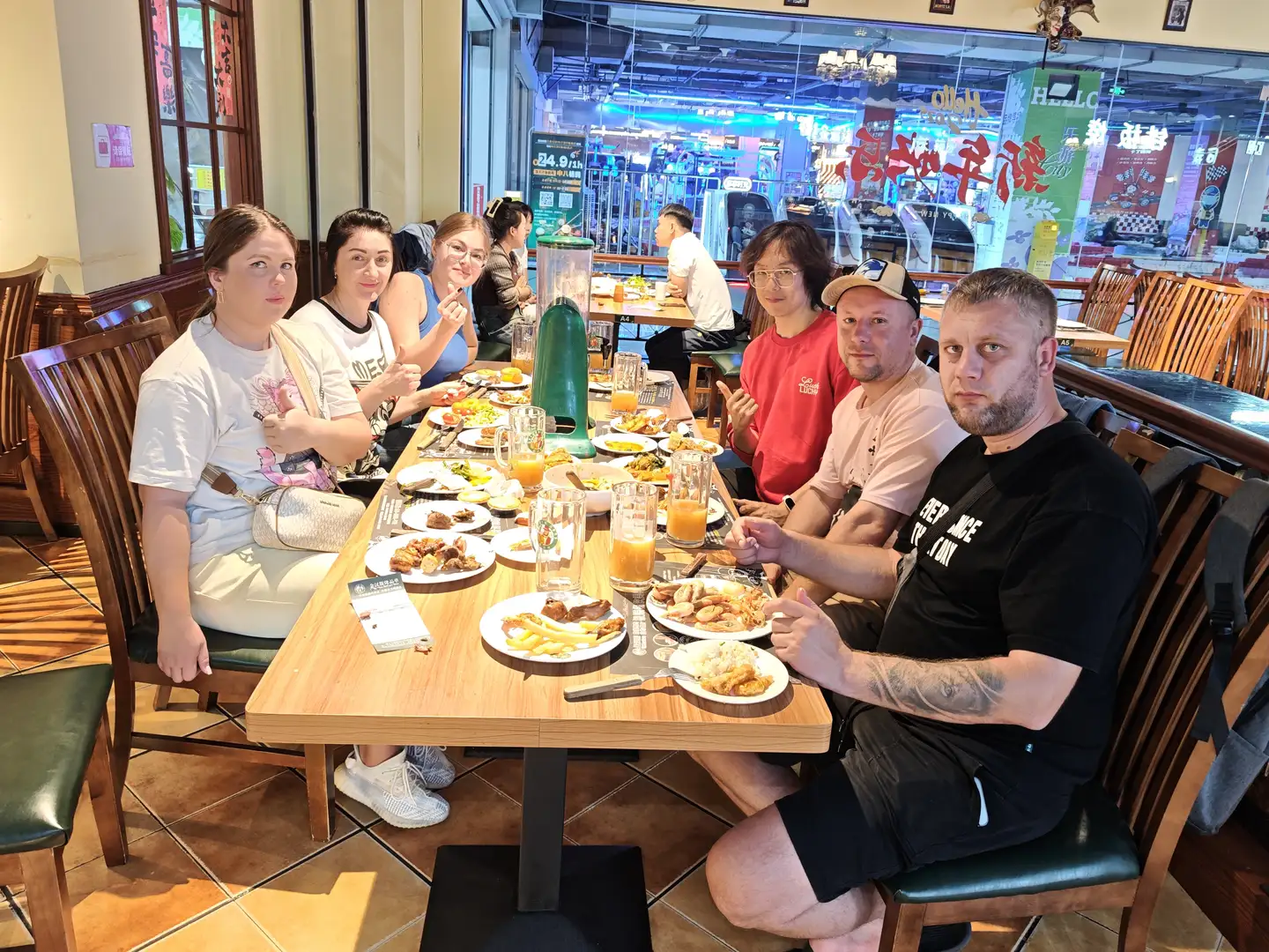 A group of people gathered around a long table in a restaurant, which is filled with food and drinks including fries, meats, and orange juice. They are dressed casually, with some smiling and others making gestures, while other customers and shop signs are visible in the background. The restaurant interior is warmly decorated with wooden tables and chairs, and large glass windows showing a brightly lit commercial street scene outside.