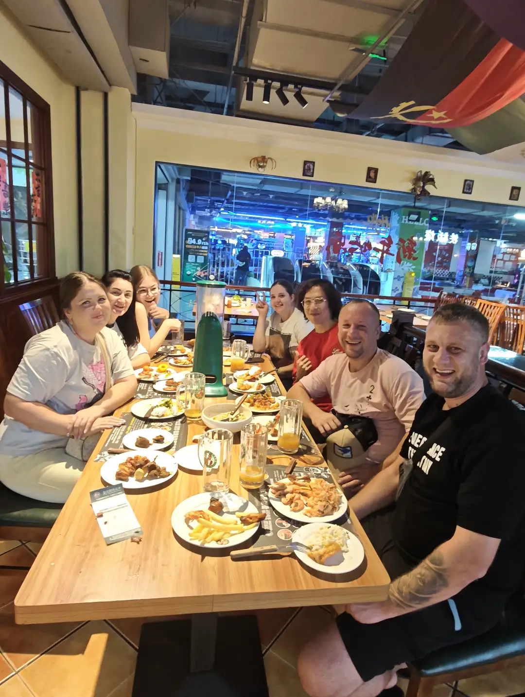 A group of people sitting around a long dining table inside a restaurant, with various dishes and beverages like fries, meats, and orange juice spread out on the table. They are in casual wear and look cheerful, with some flashing peace signs and others smiling at the camera. The restaurant is spacious and brightly lit, with red banners hanging from the ceiling, and other dining areas and shops visible in the background.