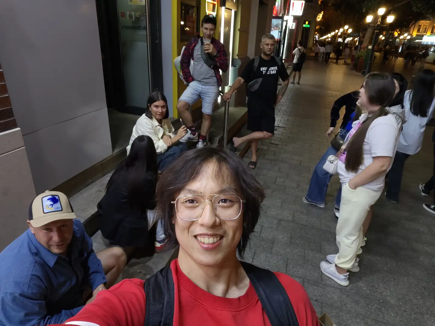 A man wearing glasses takes a selfie on a street, dressed in a red top and carrying a black backpack, with several people standing or sitting around him. The background features a city street at night, with lights illuminating pedestrians and shops, and the ground paved with stone bricks. People are in summer clothing, some chatting and others looking at their phones.