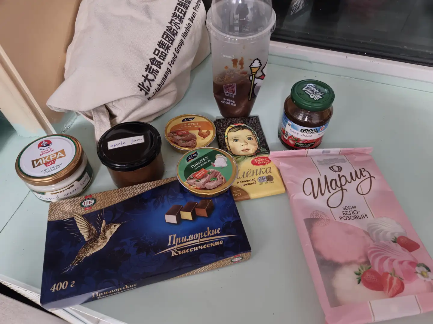 A variety of food items displayed on a tabletop, including a box of chocolates with a bird design, a bag of candies in pink packaging, several jars of jam and meat products, a cup of milk tea, and a cloth bag with Chinese characters. The chocolate box bears the Russian text 'ŠŃимоŃŃŠŗŠøŠµ ŠŠ»Š°ŃŃŠøŃŠµŃŠŗŠøŠµ', and the candy wrapper reads 'ŠŠ°ŃŠøŃŠ°' and 'ŠŠµŃŠøŃ ŠŠµŠ»Š¾-Š Š¾Š·Š¾Š²ŃŠ¹'. Jars labeled 'apple jam' and 'Global Village' jam bottles are also visible nearby.