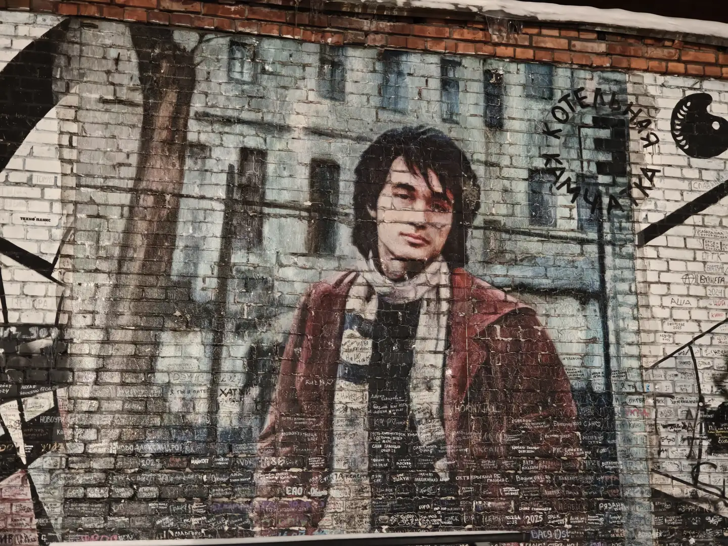“堪察加锅炉房”外部的壁画,圣彼得堡 在一面斑驳的灰白色砖墙上绘有一幅巨大的彩色壁画,画中是一位留着中长黑发、围着围巾的年轻男子半身像。壁画的上方和周围覆盖着各种黑色的手写文字和涂鸦,右侧可见圆形的字母排列。整面墙壁看起来年代久远,砖块纹理清晰可见。