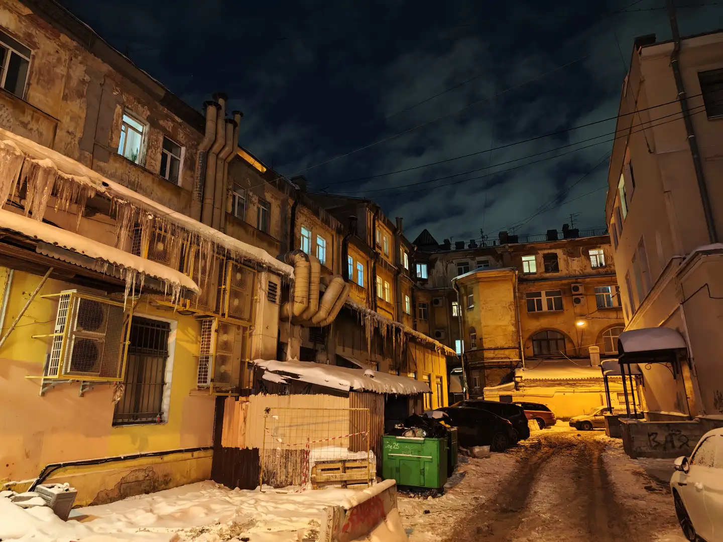 旅馆门前昏黄的街道,圣彼得堡 夜晚的庭院被积雪覆盖,左侧老旧建筑的屋檐下悬挂着长长的冰柱,窗户透出灯光。建筑外墙上有粗大的管道和空调外机,下方有一个被雪覆盖顶棚的小木屋和绿色的垃圾桶。右侧停放着几辆汽车,远处的建筑在路灯照射下呈现暖黄色,天空乌云密布。