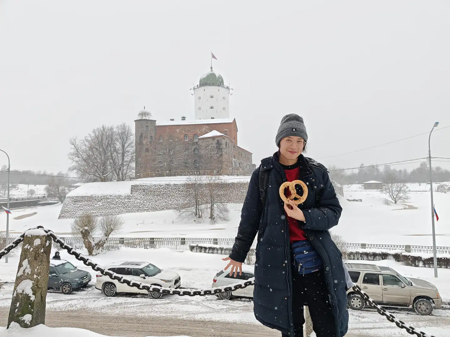 纷飞大雪笼罩的维堡城堡 背景中矗立着一座古老的石砌城堡,拥有红砖墙体和白色的圆形塔楼,塔顶飘扬着旗帜,周围覆盖着厚厚的积雪。前景中站着一位戴着眼镜和灰色针织帽的人,身穿深蓝色长款羽绒服,手中拿着一个巨大的椒盐卷饼形状的面包。人物身后是一排停放的汽车和一条被铁链围起来的道路,天空中飘着雪花,地面和树木都被白雪覆盖。