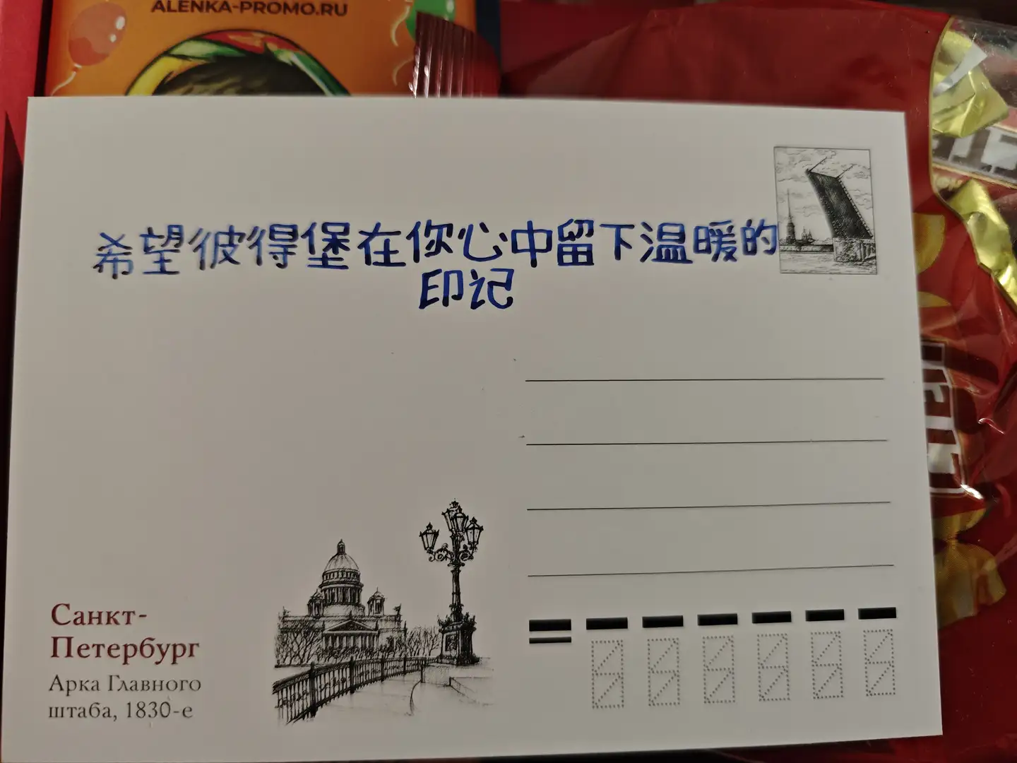 热尼亚送给我的明信片 明信片背面的上方用蓝色墨水手写着中文“希望彼得堡在你心中留下温暖的印记”,右侧有一幅小桥的素描画。左下角印有黑色的“Санкт-Петербург”(圣彼得堡)字样以及一座圆顶建筑的素描图,旁边还有路灯图案。右侧留有四行横线用于书写地址,下方有七个虚线方框组成的邮政编码区域,背景隐约可见红色的糖果包装纸。