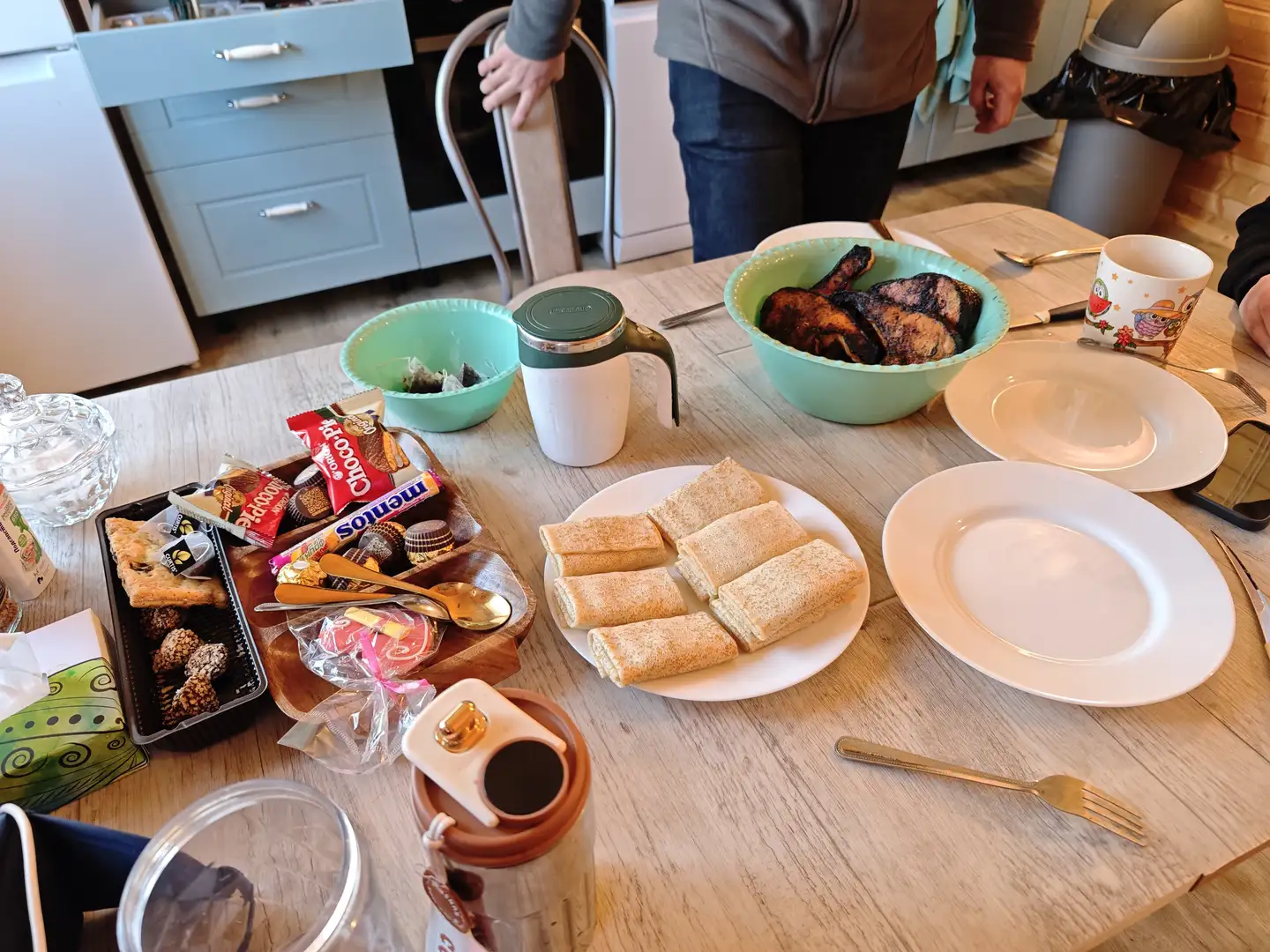 科利亚家的餐桌 一张浅色木纹桌面上摆放着各种食物和餐具,包括一盘切成长条状的三明治或面包卷。桌子左侧有一个木制托盘装着巧克力和其他零食,旁边是一个绿色碗里泡着茶包和一个保温杯,右上方还有一个绿色碗装着看起来像烤肉的食物。桌子上还散落着白色的盘子、叉子、勺子和一个带有猫头鹰图案的马克杯,背景中有穿着牛仔裤的人站在厨房柜子旁。