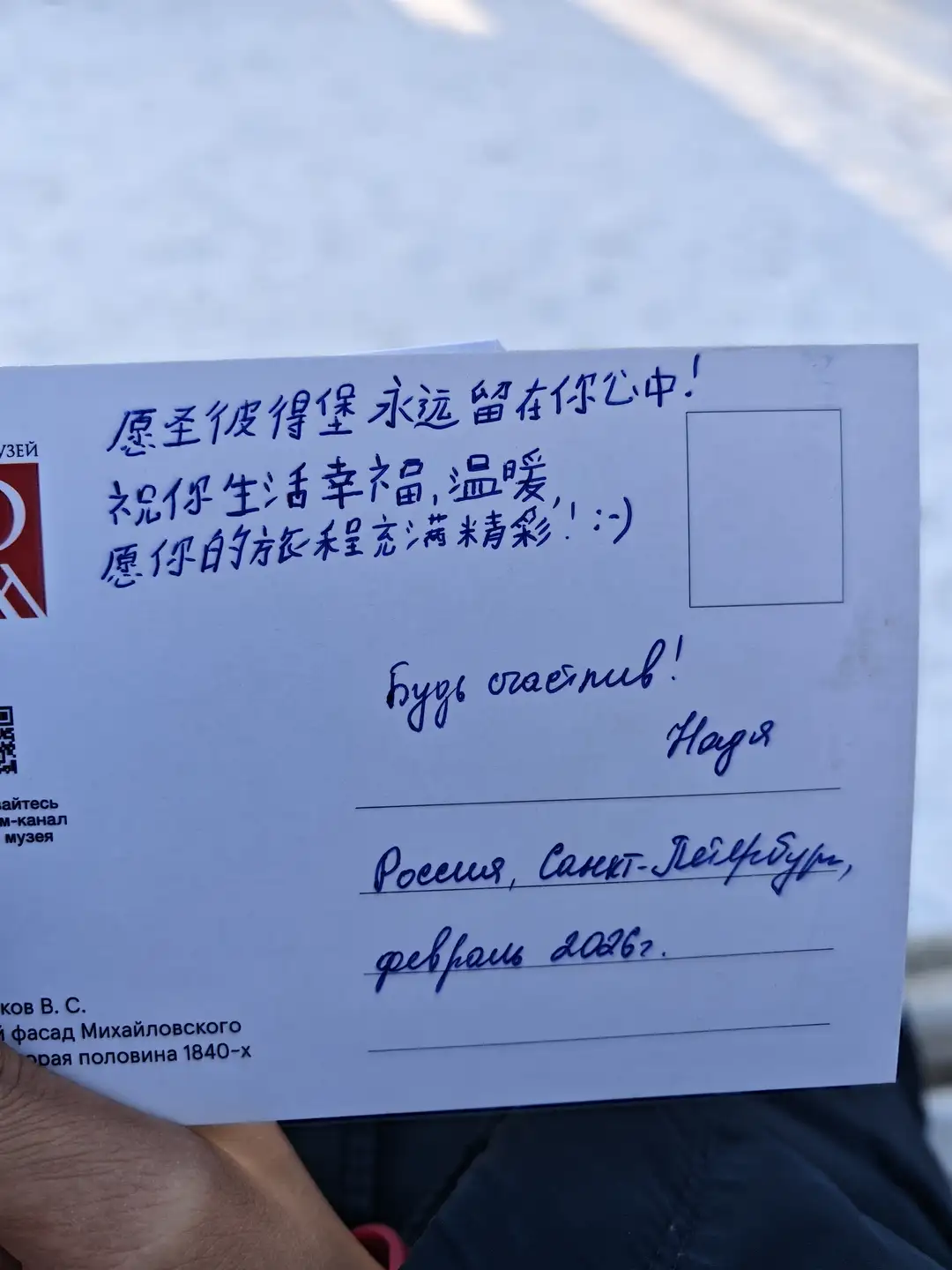 娜佳送给我的明信片 明信片背面上方用蓝色墨水手写着中文祝福语“愿圣彼得堡永远留在你心中!祝你生活幸福温暖,愿你的旅程充满精彩!:- )”。中间部分用草写体写着俄语“Будь счастлив!”(祝幸福!)并署名“Надя”(娜佳)。下方地址栏手写了“Россия, Санкт-Петербург, февраль 2022.”(俄罗斯,圣彼得堡,2022年2月),左上角有部分红色标志和二维码。