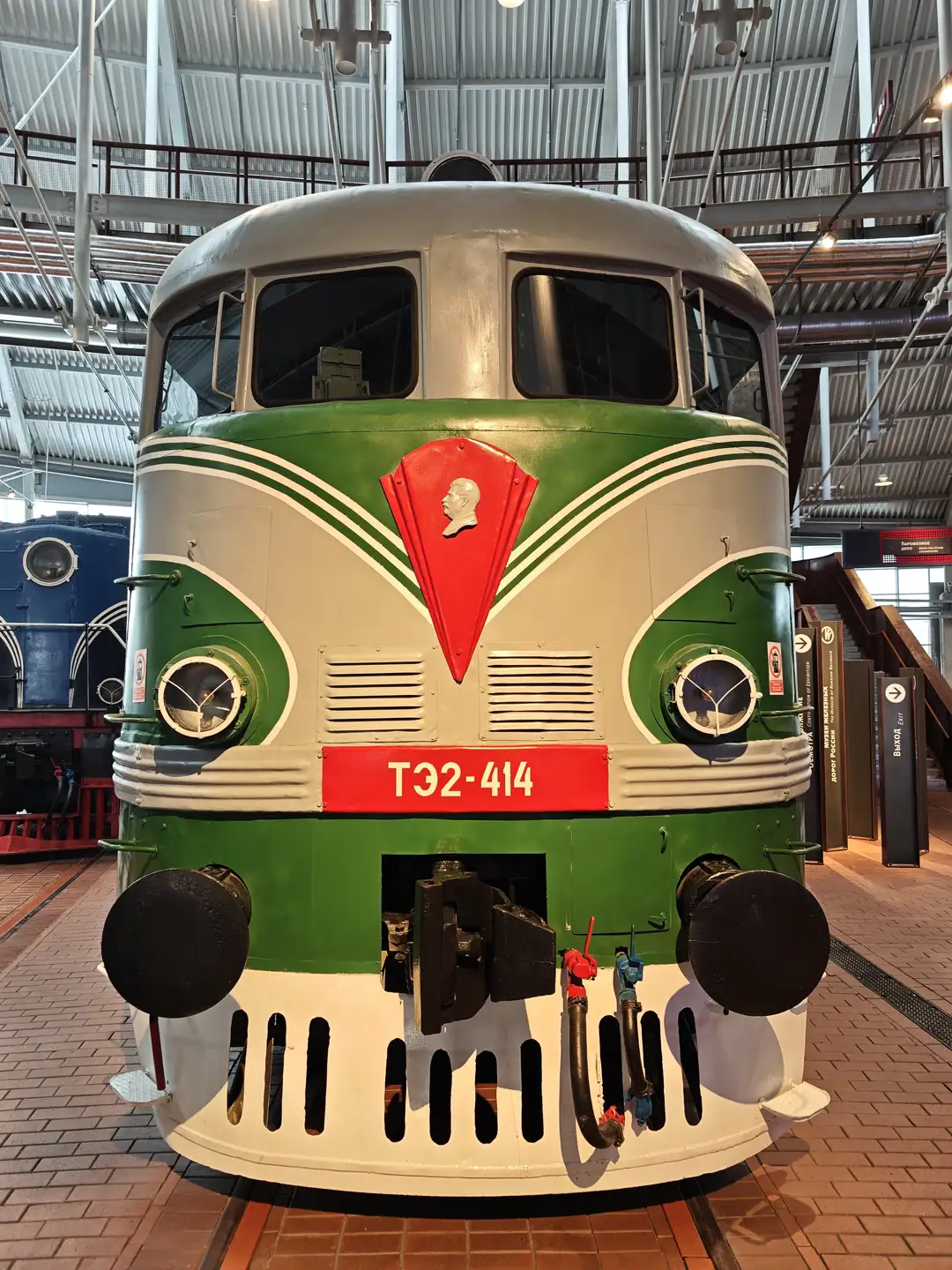 ТЭ2-414 型机车,铁路博物馆,圣彼得堡 一辆绿白相间的机车正面朝向镜头,车头中央有一个红色的盾形标志,上面印有列宁的侧面浮雕。车头下方的红色长条区域写着白色文字“ТЭ2-414”。两侧圆形的车灯和底部的黑色缓冲器清晰可见,背景是工业风格的展厅屋顶。