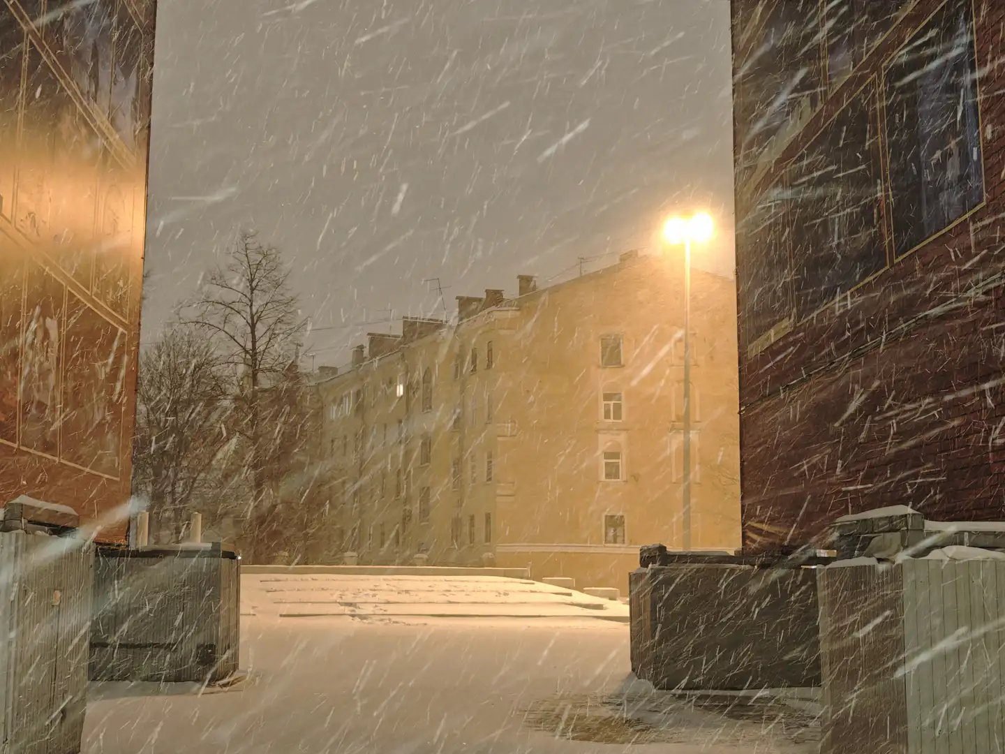 暴风雪,马林斯基剧院音乐厅,圣彼得堡 大雪纷飞,密集的白色雪花在画面中形成斜线,覆盖了整个视野。画面两侧是深红色的砖墙建筑,中间透过空隙可以看到远处一栋黄色的多层公寓楼。一盏明亮的路灯矗立在远处建筑旁,发出暖黄色的光芒,地面和前景的物体上都积满了厚厚的雪。