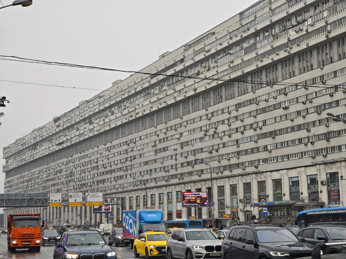 原子科学家大楼,莫斯科 一座极长的大型多层建筑横向延伸,其立面布满了密密麻麻的窗户和悬挂的空调外机。建筑底层是一排排临街的商铺入口,上方则是重复排列的办公或居住空间。前景是拥堵的城市街道,行驶着橙色的卡车、印有“OZON”字样的蓝色货车、出租车以及私家车。