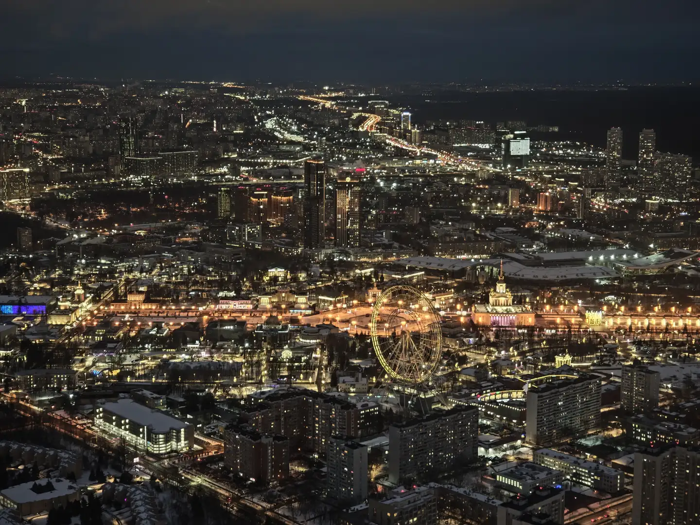 夜幕下的莫斯科,奥斯坦金电视塔,莫斯科 一张城市夜景的俯瞰图,无数灯光点缀着密集的建筑群。画面右下方有一个巨大的摩天轮,其轮廓被灯光照亮,旁边是一座带有尖顶的古典建筑。远处可以看到蜿蜒的街道灯光带和黑暗的水域,高楼大厦在夜色中耸立。
