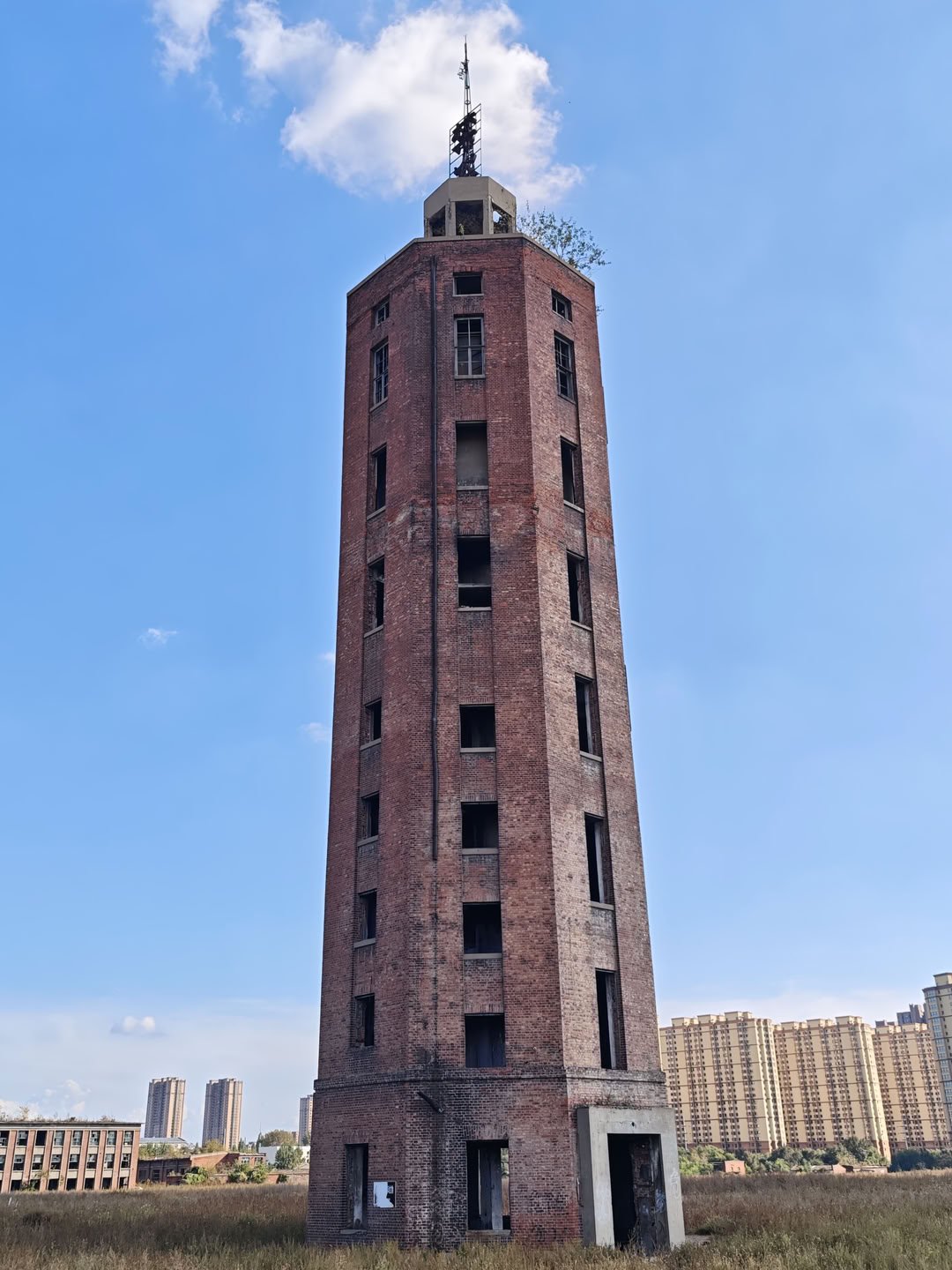 一座高耸的红砖塔楼占据画面主体,塔身分布着多层长方形窗户。塔顶设有金属框架结构,周围生长着少量树木。远处背景中可见几栋米黄色的高层住宅楼,天空中有白云。