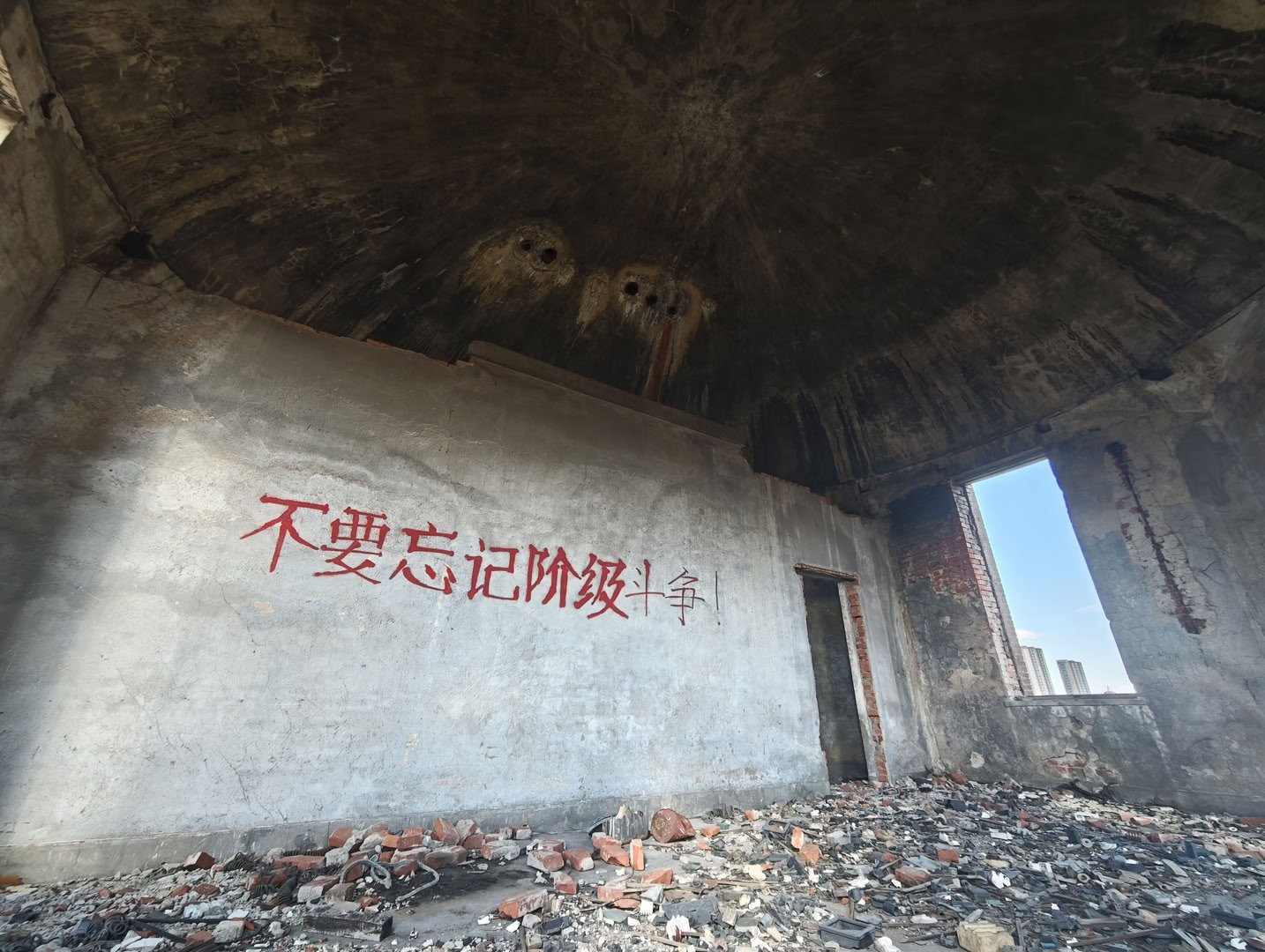 这是一个内部呈圆顶结构的废弃房间,地面堆满了碎砖和瓦砾。正对面的灰色墙壁上用红色颜料写着“不要忘记阶级斗争!”的大字。右侧有一个长方形门洞和窗户,透过窗户可见外面的蓝天和建筑。