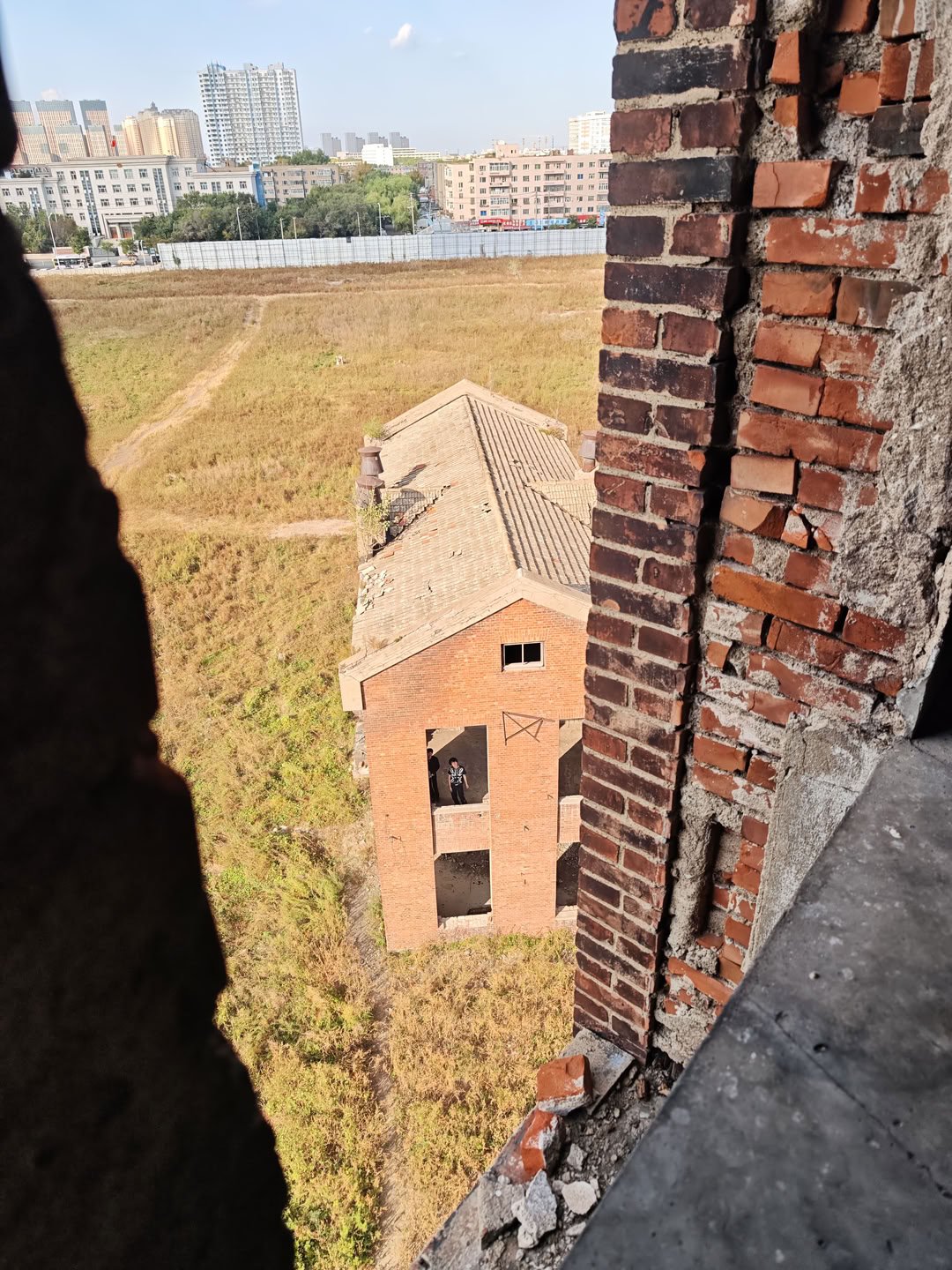 右侧粗糙的红砖墙体和混凝土边缘构成了前景框架,左侧被巨大的黑色阴影遮挡,视线向下延伸至一片荒草丛生的空地。空地上矗立着一座红砖建筑,其灰色屋顶略显破旧,建筑正面的矩形开口处站着两个人影。远处可以看到城市的天际线,包括高层住宅楼、低矮的建筑群以及蓝色的施工围挡。